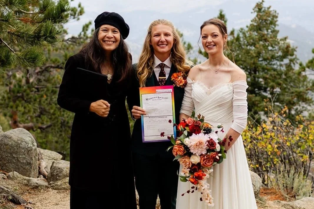 Officiant Lisa and two brides smile as one bride holds up their rainbow-bordered marriage license.