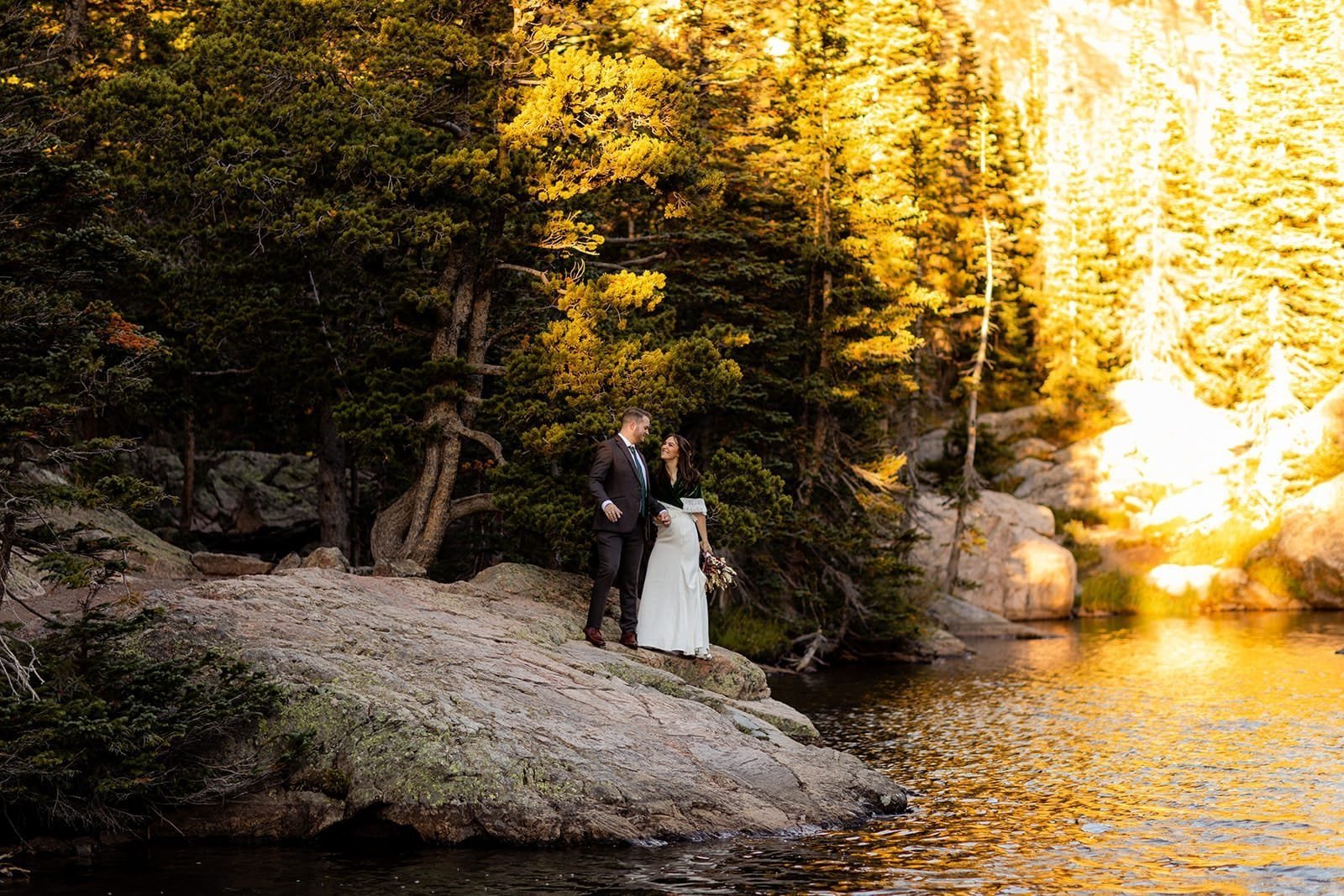 A bride and groom hold hands while standing on a granite rock that juts out into an alpine lake with fir trees.