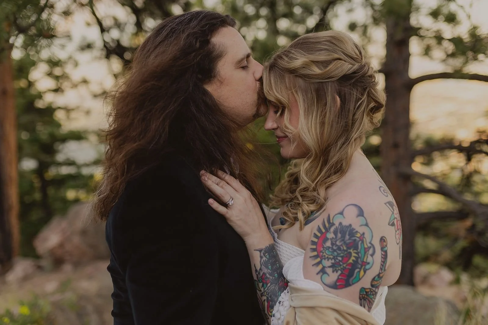 A groom kisses his tattooed bride on the forehead while she gently places her hand on his chest.