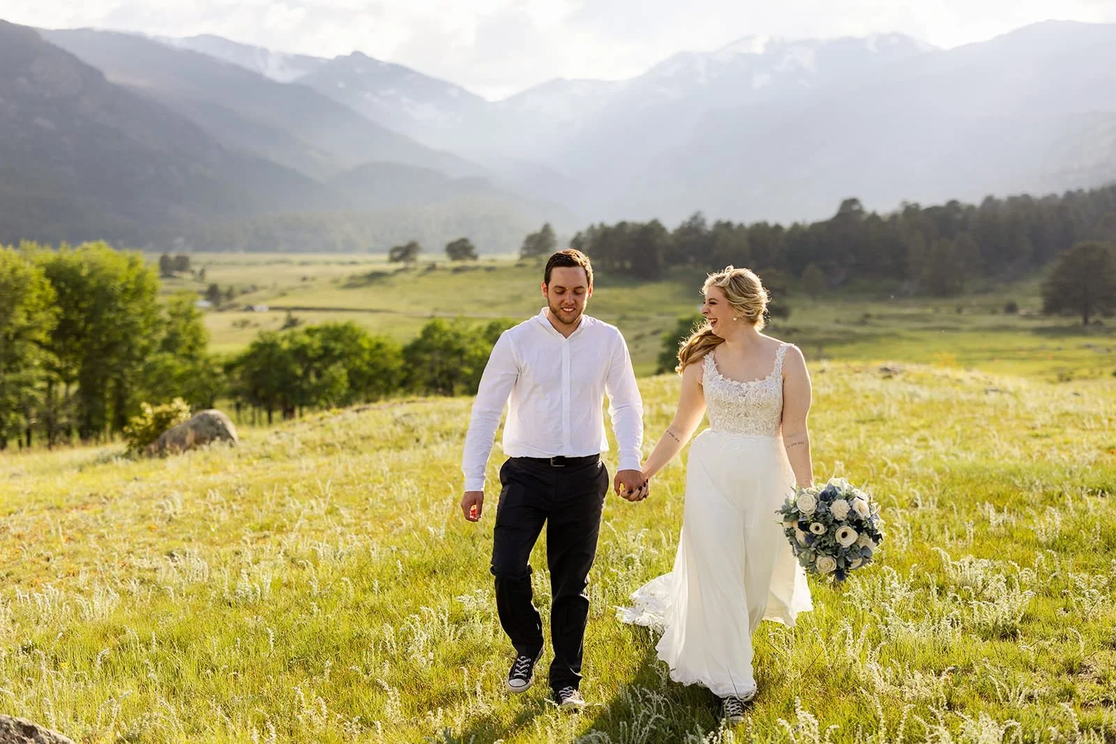 An eloping bride and groom hold hands and smile while walking through a bright green meadow with a misty mountain backdrop.