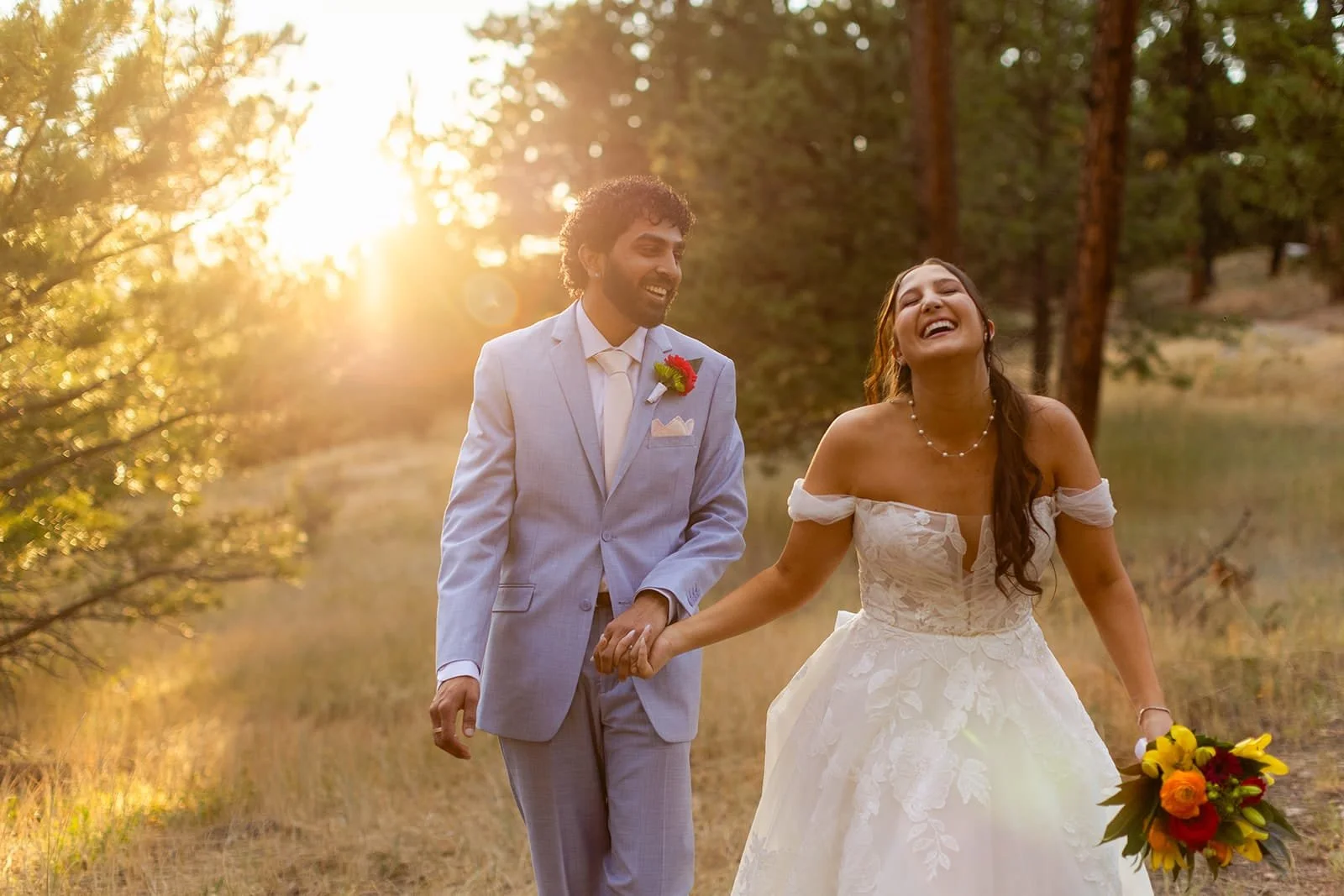 A bride and groom laughing as they walk along a forest path during sunset.