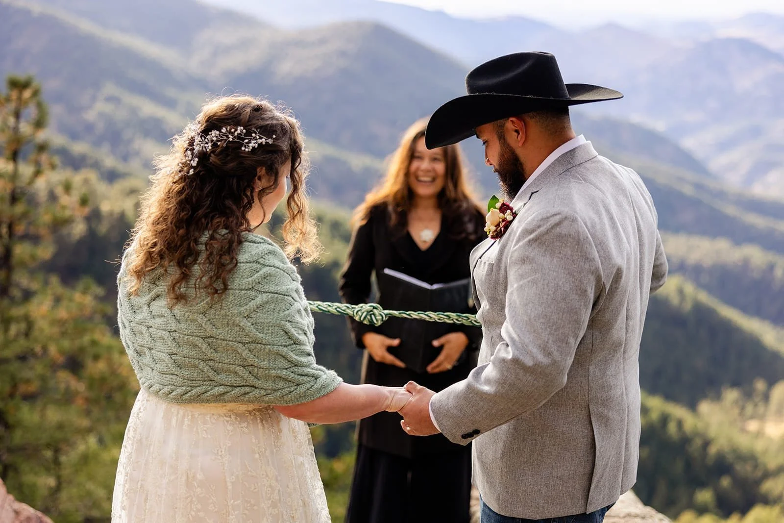An eloping bride and groom tie a hand fasting knot while Officiant Lisa smiles with a rolling mountain hill backdrop.