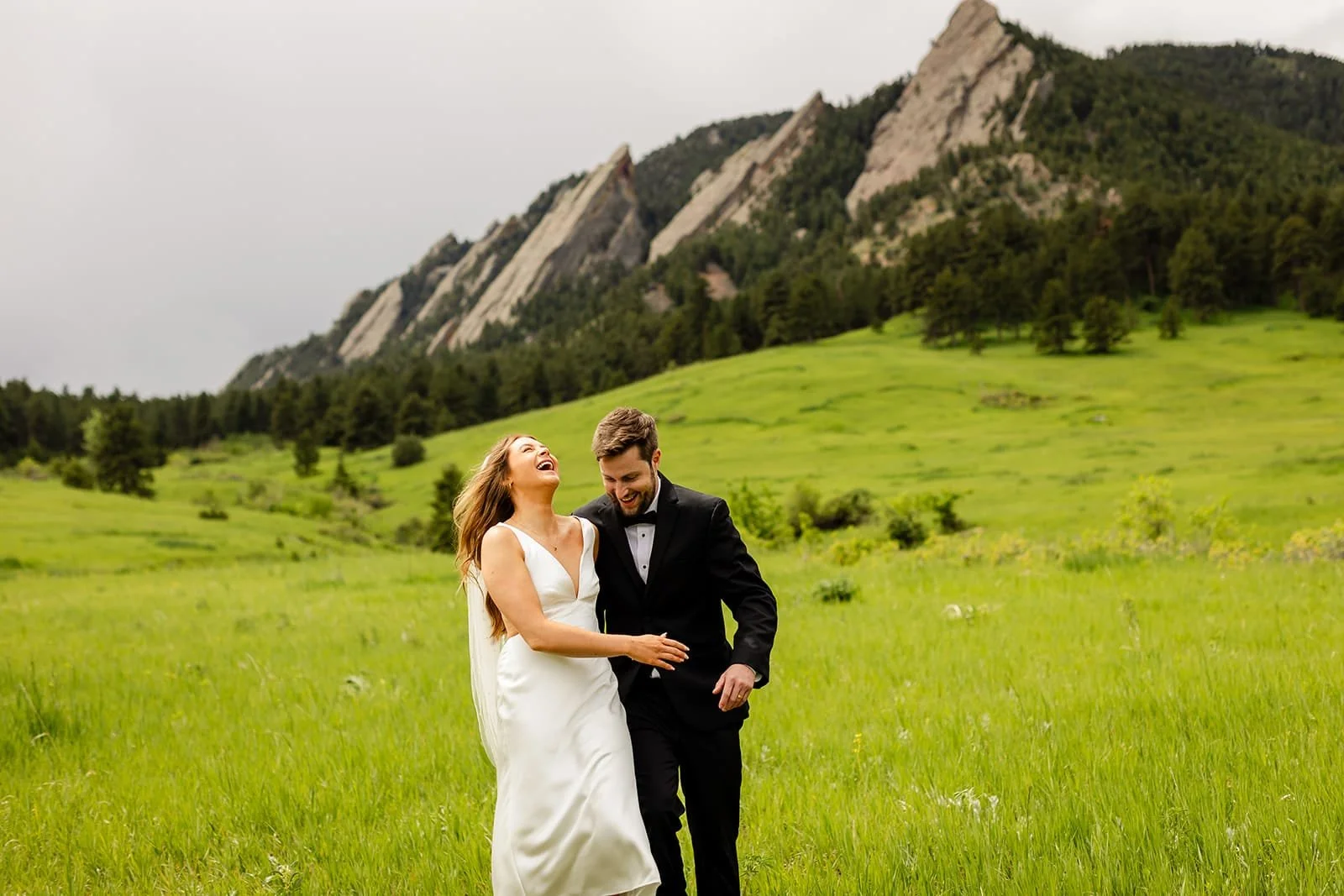 A just-married eloping couple dance in a bright green meadow while laughing and with large rock formations behind them.