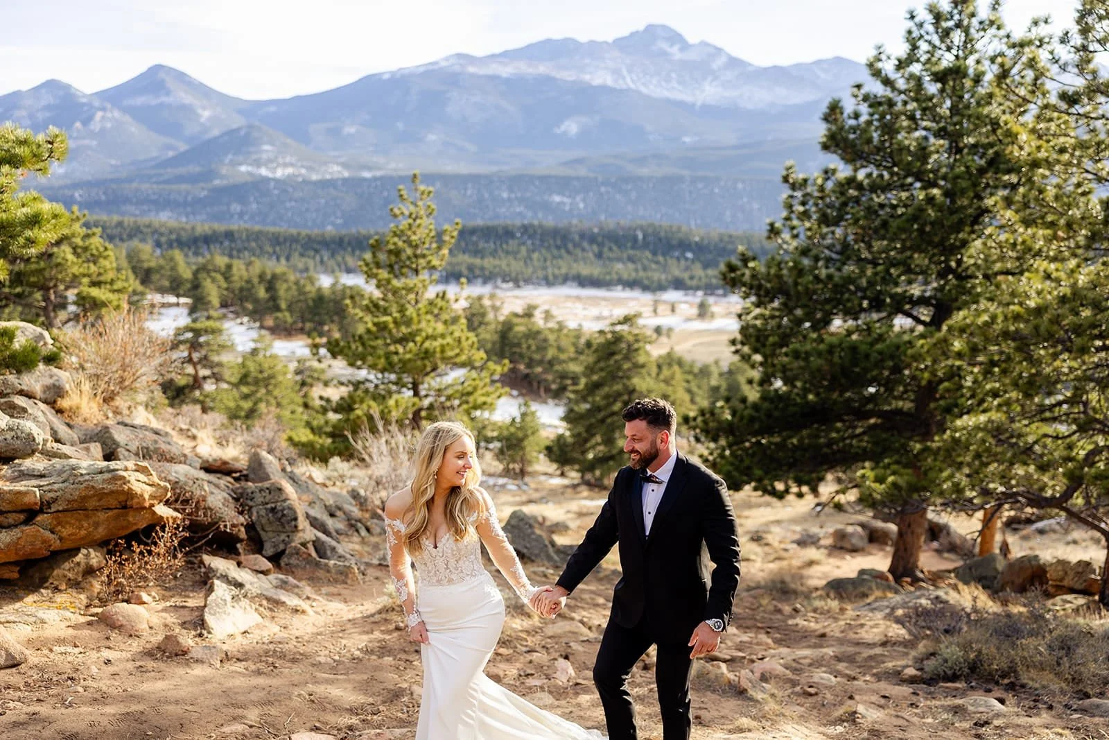 An eloping bride and groom walk playfully among trees with a mountain backdrop.