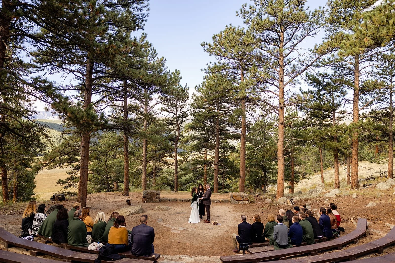 Officiant Lisa guides a micro wedding ceremony in a forest amphitheater with 26 guests seated on wooden benches.