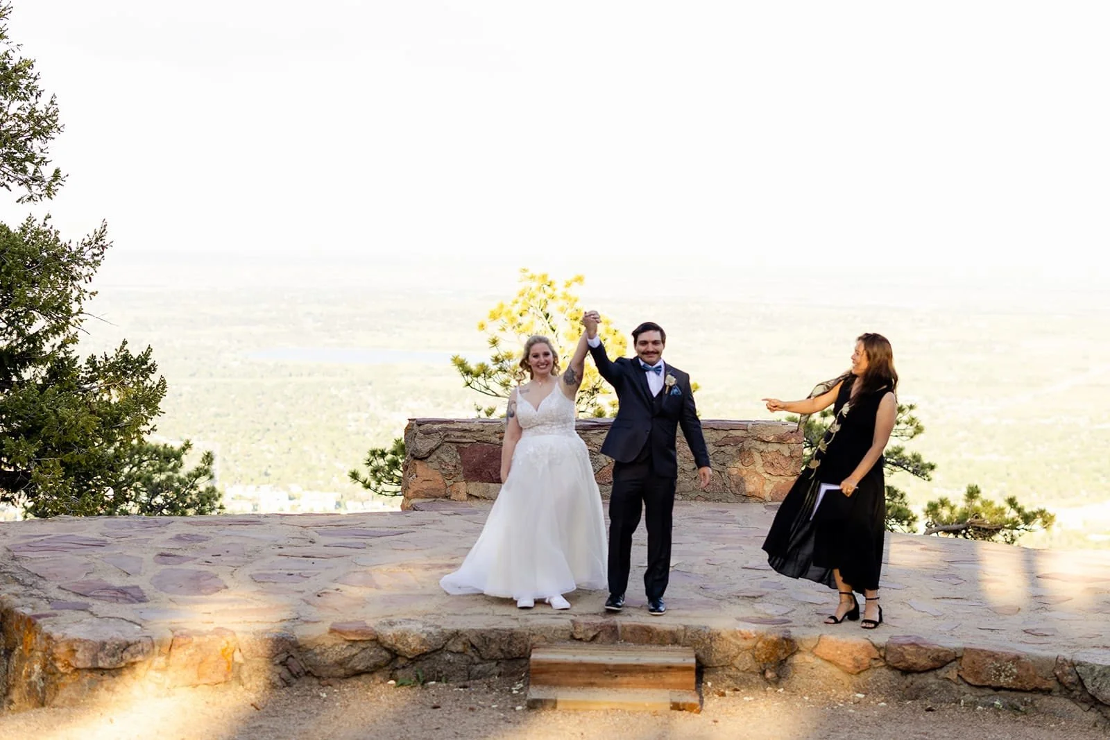 A bride and groom raise hands in triumph as Officiant Lisa declares them married on the stone stage at sunset.