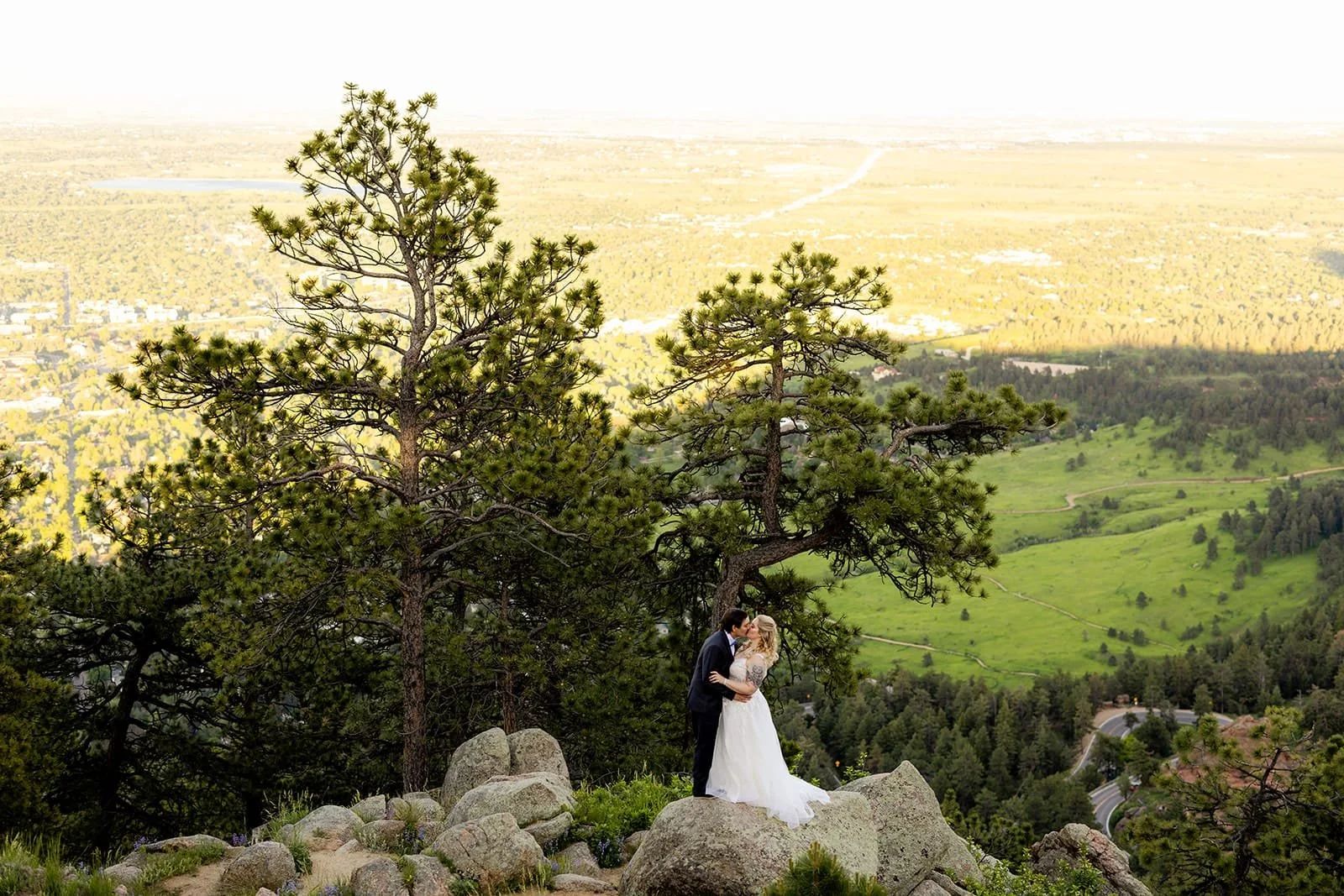 A wide shot of pine trees and Colorado plains with a couple kissing on granite rocks at the edge of the mountain.