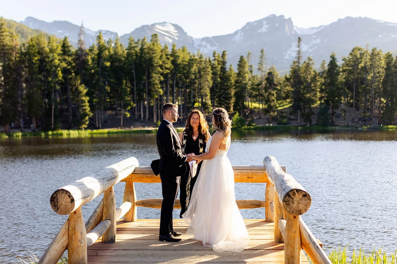 Officiant Lisa guides an elopement ceremony on a wooden dock at an alpine lake with sunset forest and mountain backdrop while the couple look into each others' eyes.