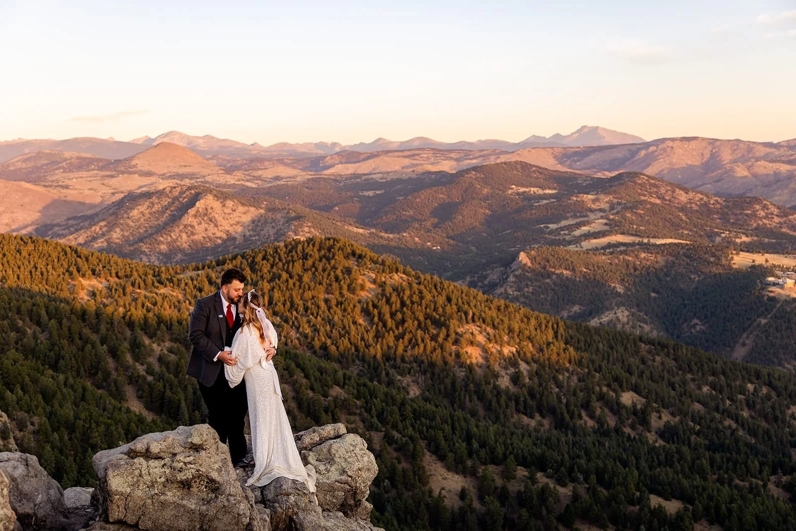 A just-married eloping couple hugs while standing on rocks on the edge of the mountain with a vast rolling mountain sunset backdrop.