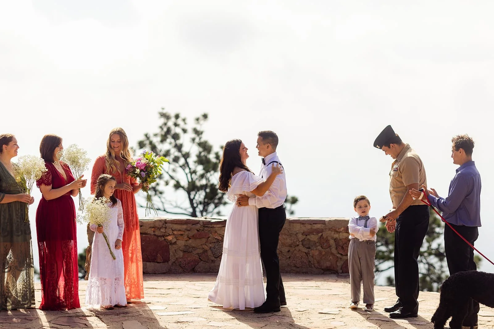 A bride and groom hug during their micro wedding ceremony while bridesmaids, groomsmen and a ring bearer clap and look on.