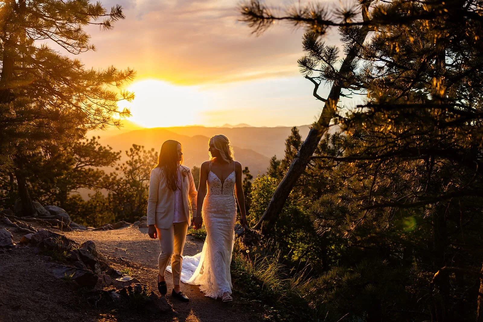 Two brides walk while holding hands with a mountain backdrop and setting sun.