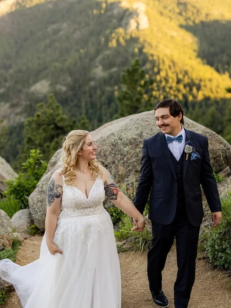 A couple hold hands while walking amidst granite rocks and trees.