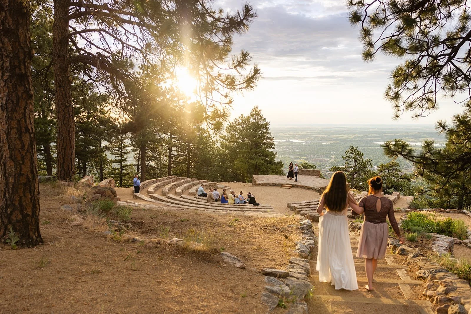 A bride walks her wedding processional with her mother down the stairs into Sunrise Amphitheater as the rising sun shines, guests watch and her groom waits on the stage.