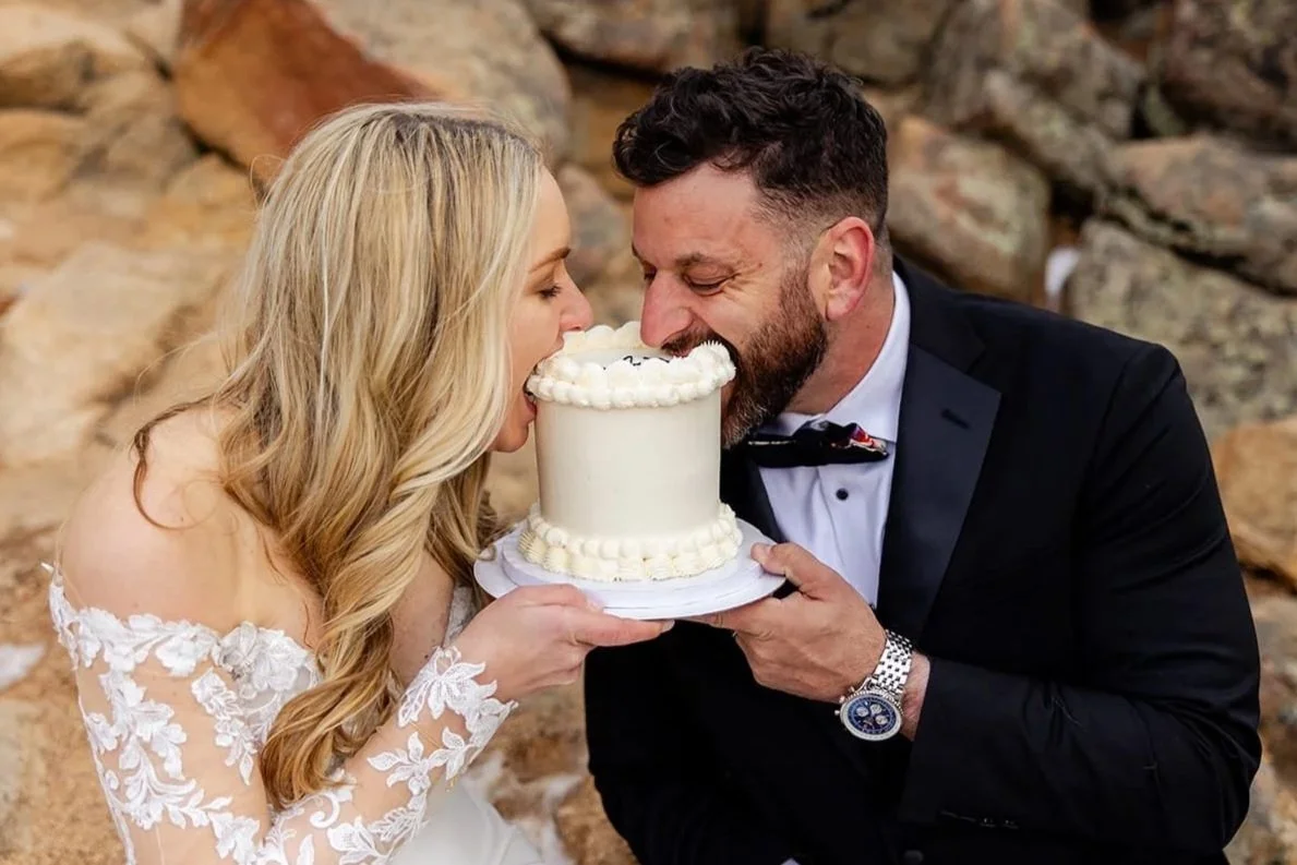 A just-eloped bride and groom hold their small wedding cake together while both playfully biting into it at the same time.