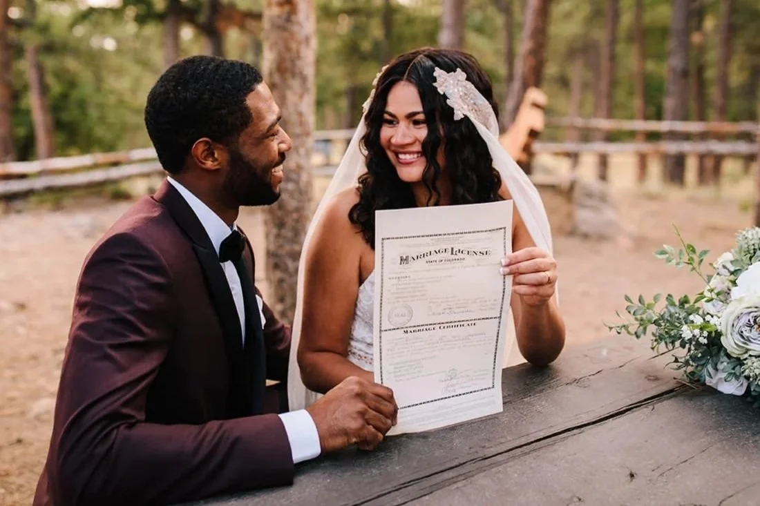 A bride and groom smile at each other as they hold up their just-signed marriage license while sitting at a picnic table in the forest.