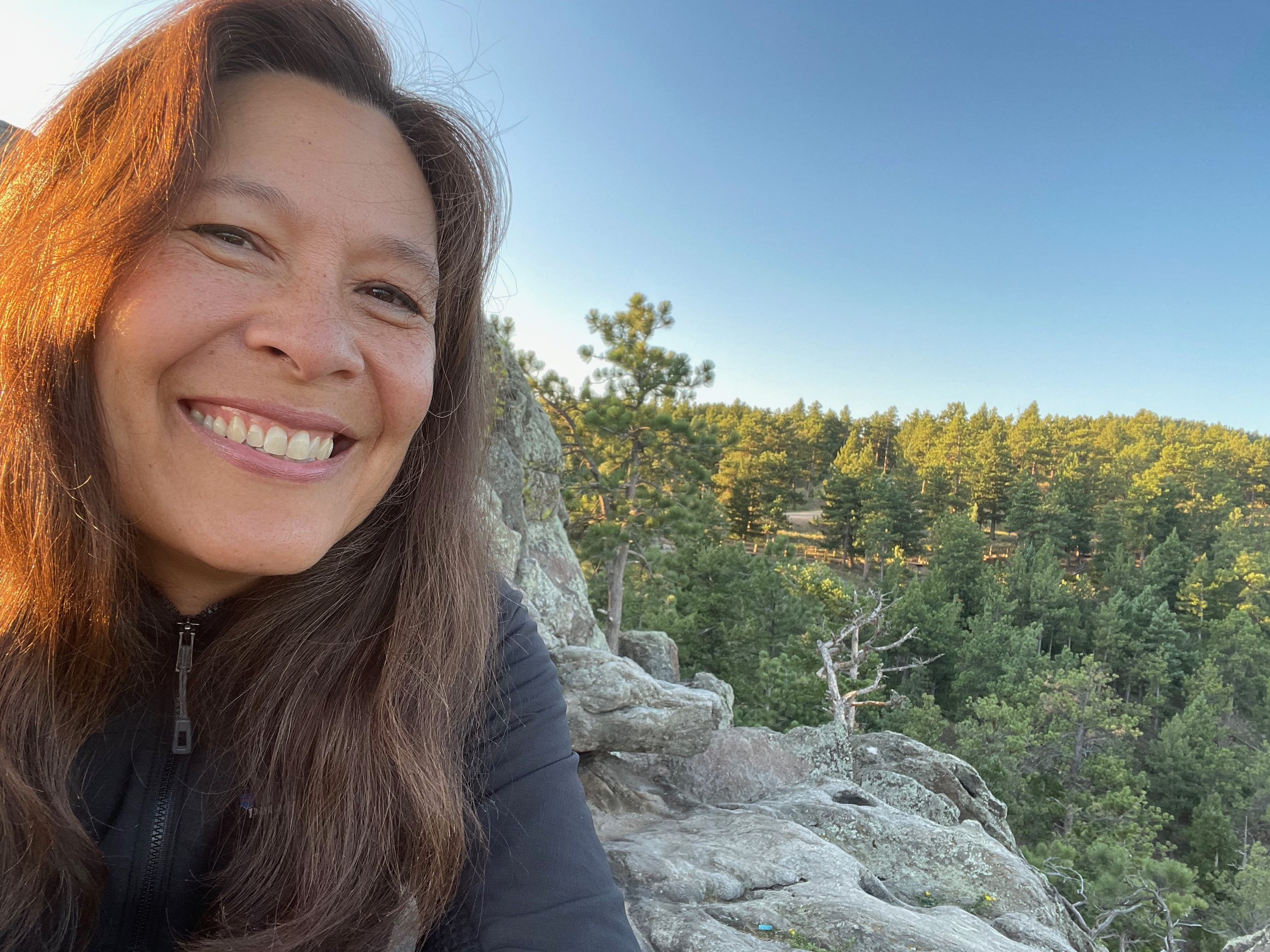 Officiant Lisa smiles while sitting on granite rocks as the sun rises with a forest backdrop.