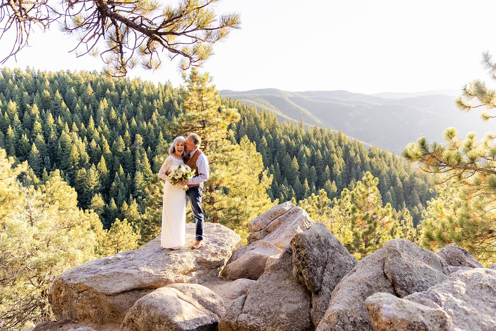 A groom kisses his bride who has a huge funny smile and they are standing on a big rock with a forested rolling hill backdrop.