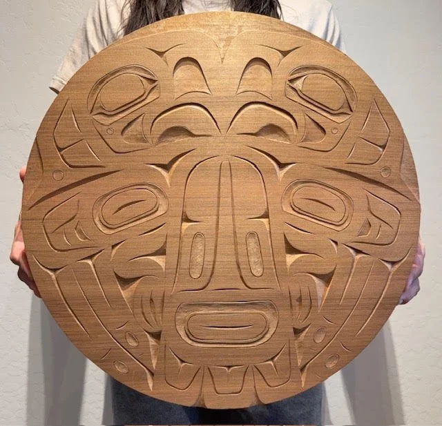 Person holding a circular carved panel in the Tlingit style.