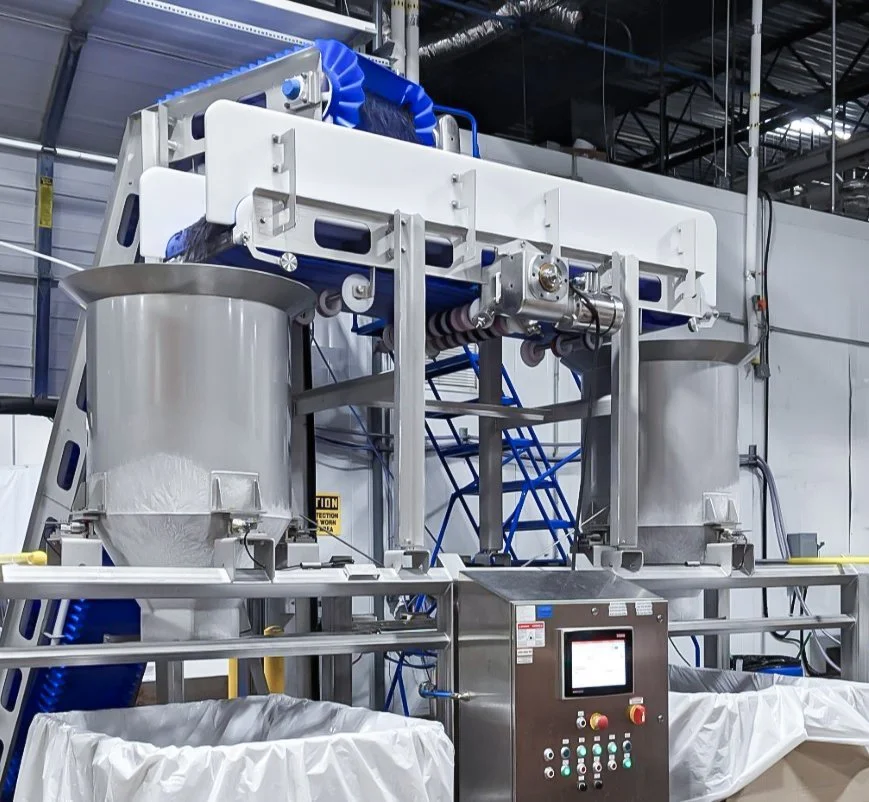 A weigh hopper with stainless steel tanks, a control panel, and blue conveyor rollers in a factory setting used for strawberries.