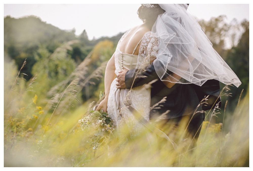 Couple embrace amidst tall grass and trees at Brickworks on their summer wedding.