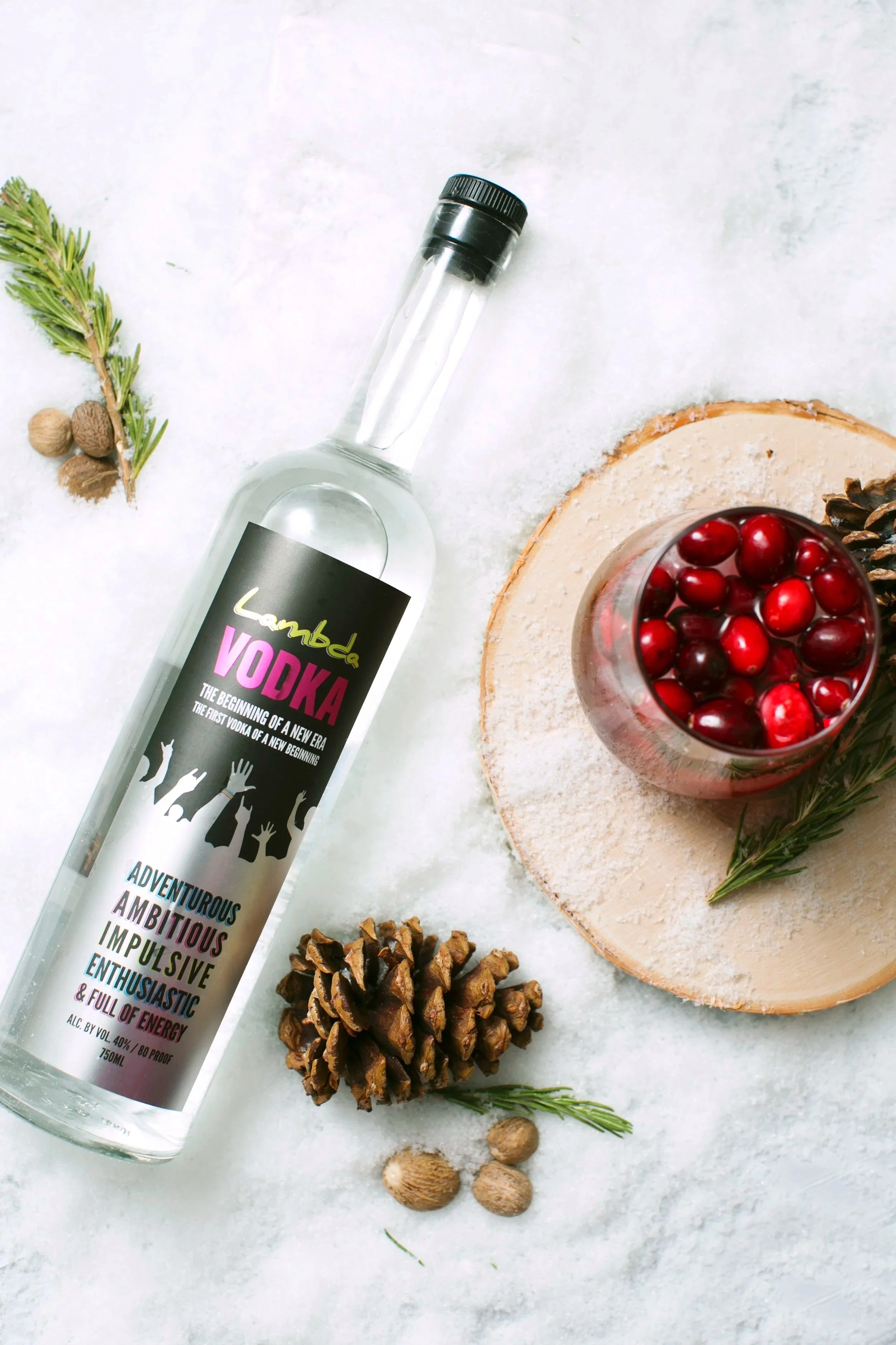 Bottle of vodka with a label that says 'Lambda VODKA,' a glass filled with cranberries, pinecones, and herbs on a white surface.