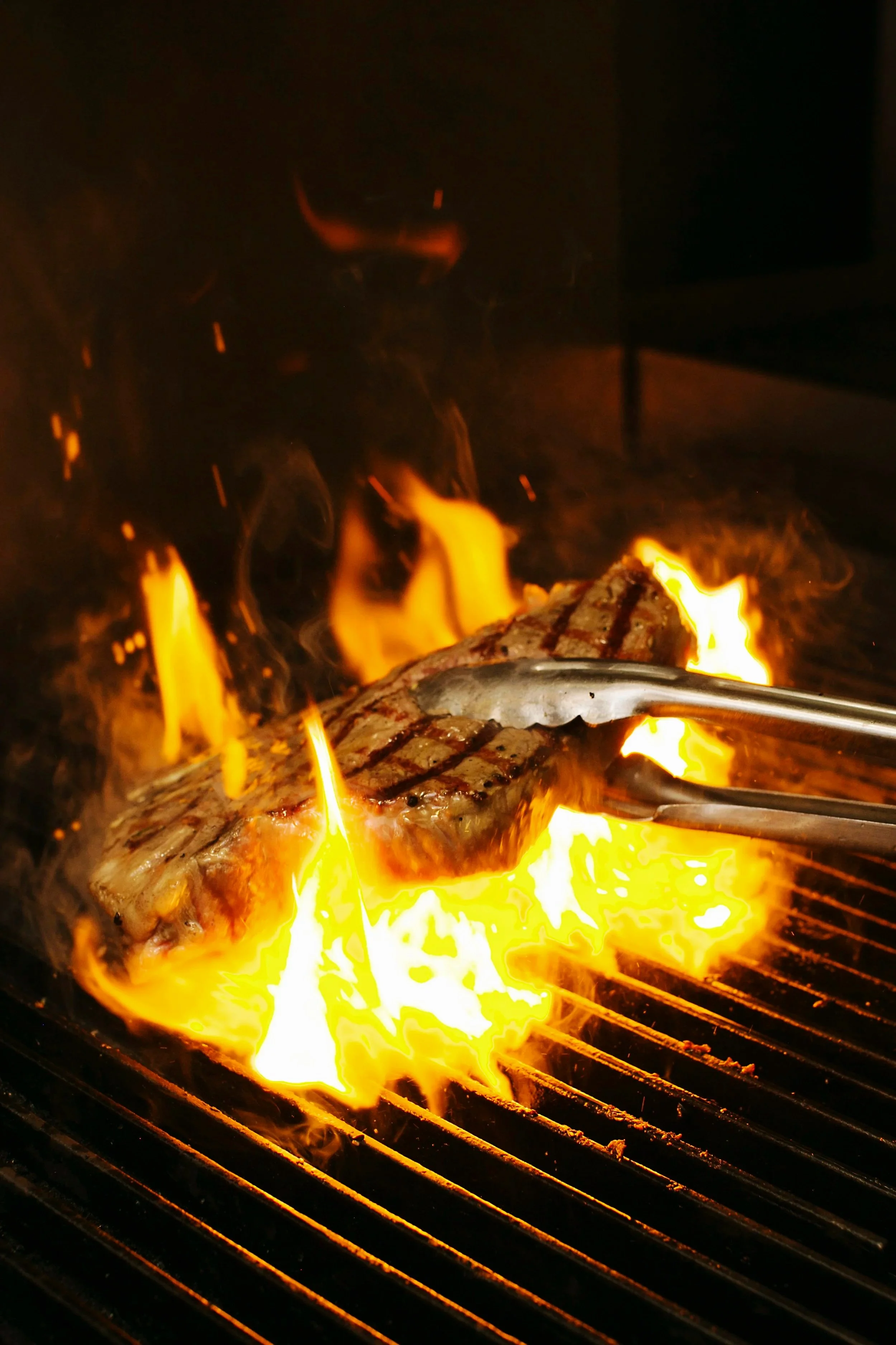 A piece of steak cooking on a hot grill with flames, being held with tongs.