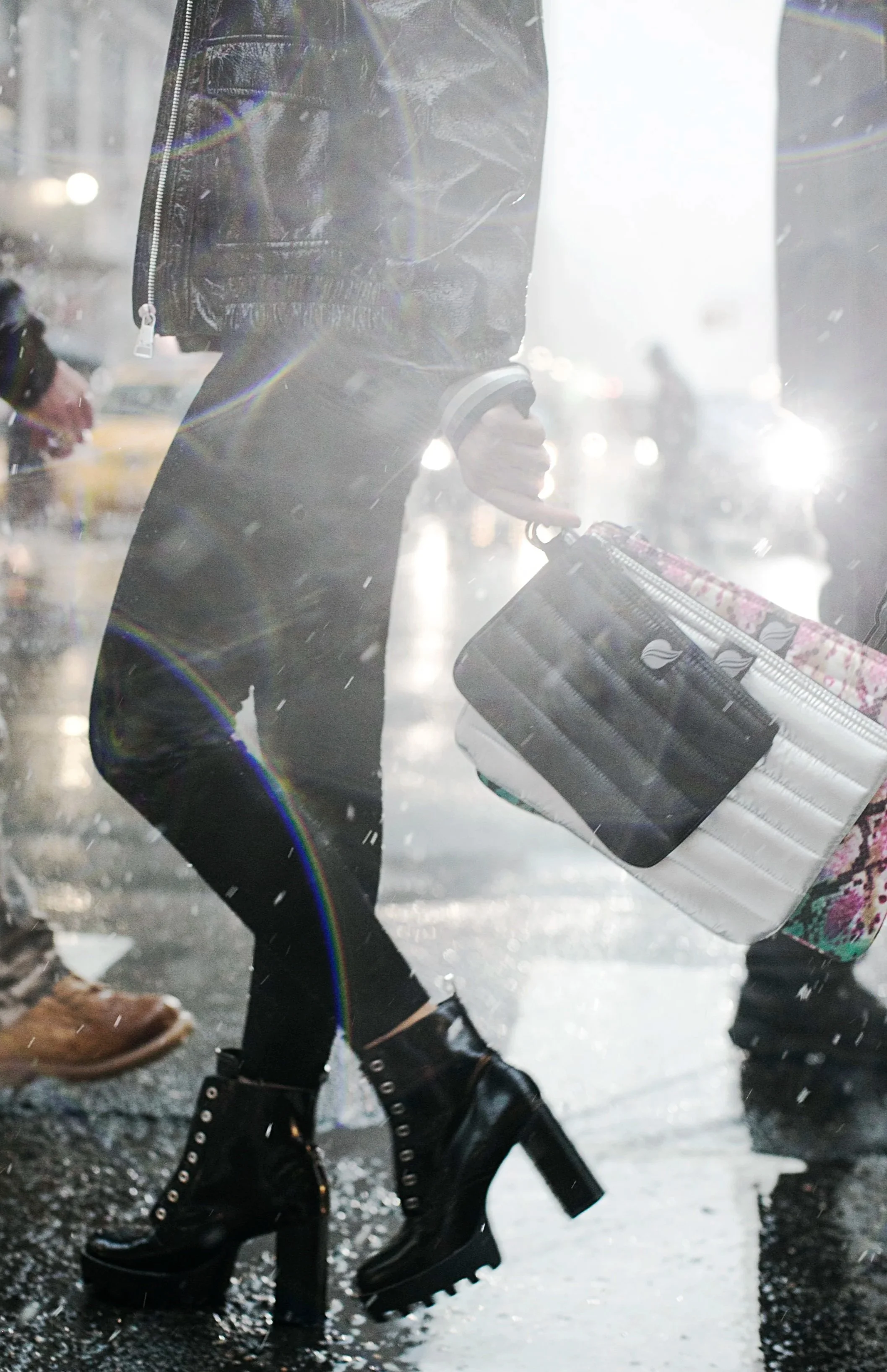 Close-up of a woman walking on a rainy city street, wearing black high-heeled boots with laces, carrying a patterned purse and a paper bag, with sunlight and raindrops visible.