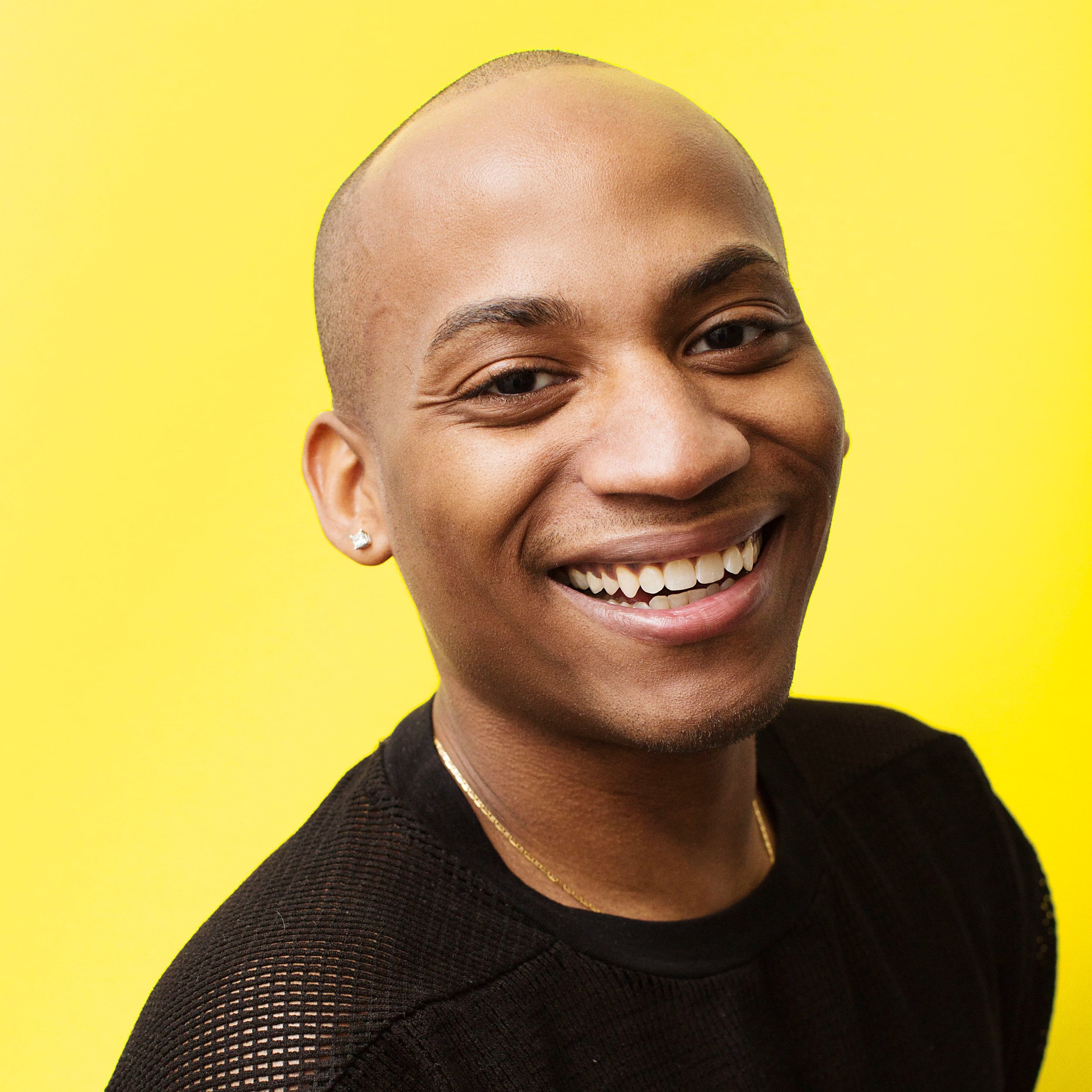 Smiling young person with shaved head, wearing earrings, a gold chain, and a black shirt, against yellow background.