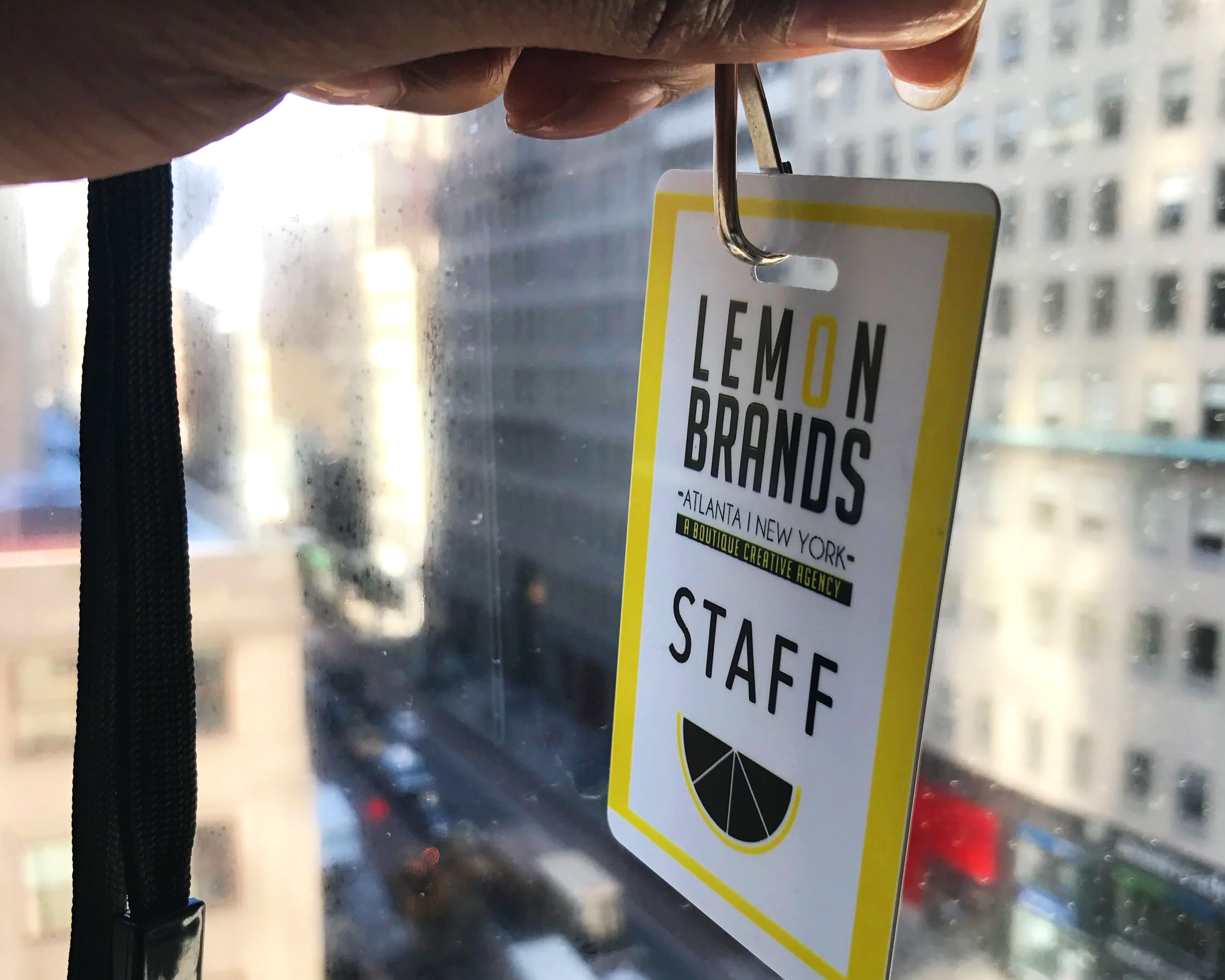A staff badge hanging on a window hook with a cityscape view in the background. The badge is from Lemon Brands, a creative agency, and indicates the individual is staff.
