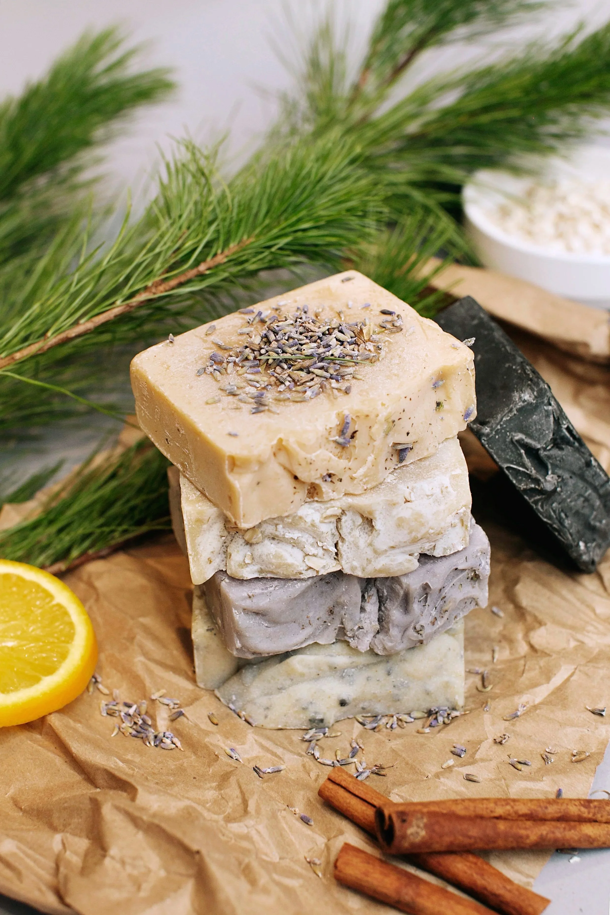 Stacked handmade soaps with lavender on top, surrounded by rosemary sprigs, a lemon slice, cinnamon sticks, and a small bowl of white powder on crumpled brown paper.