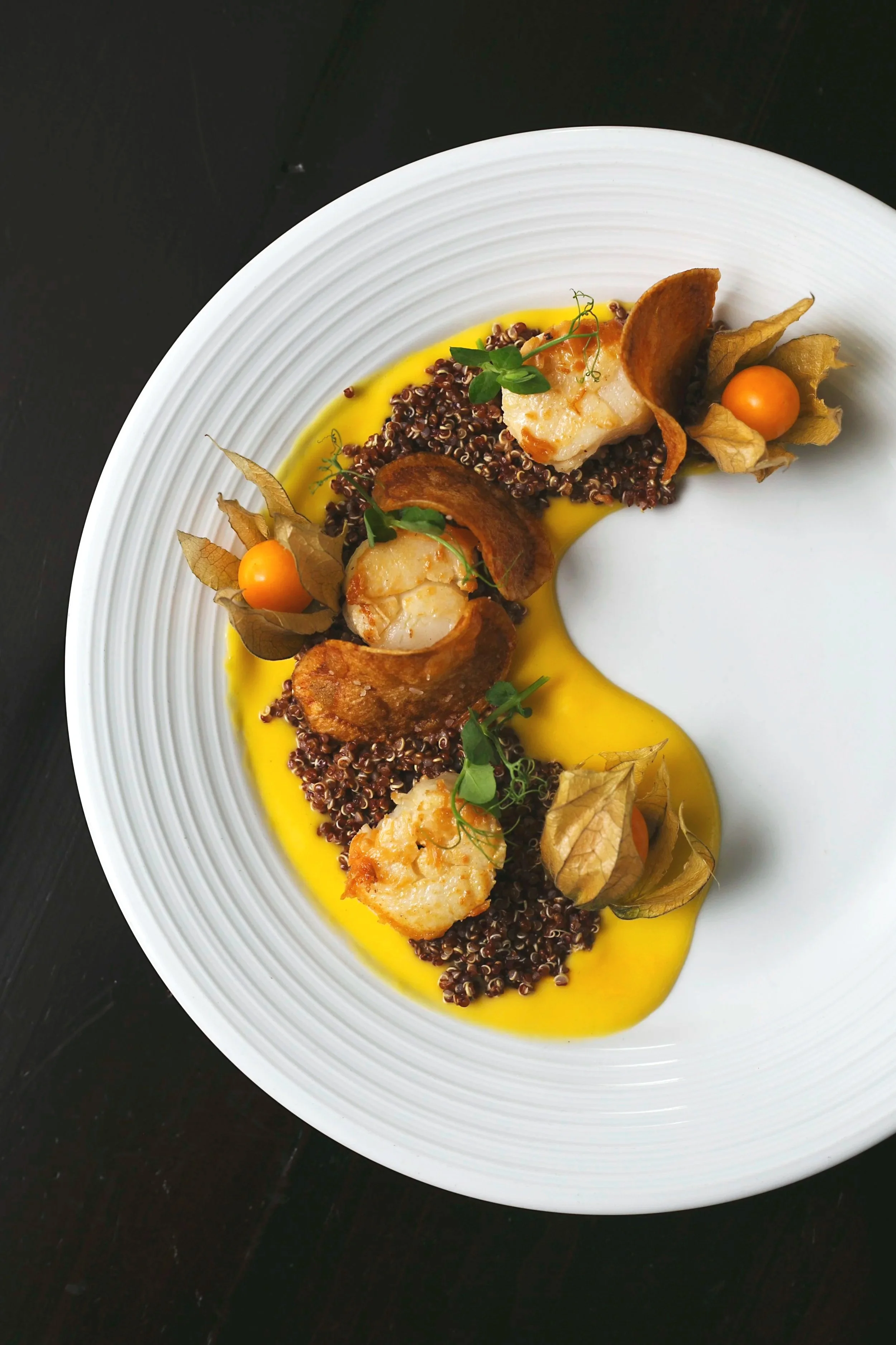 A white plate with cooked shrimp, fresh herbs, quinoa, and sauce garnished with halved physalis fruits and microgreens.