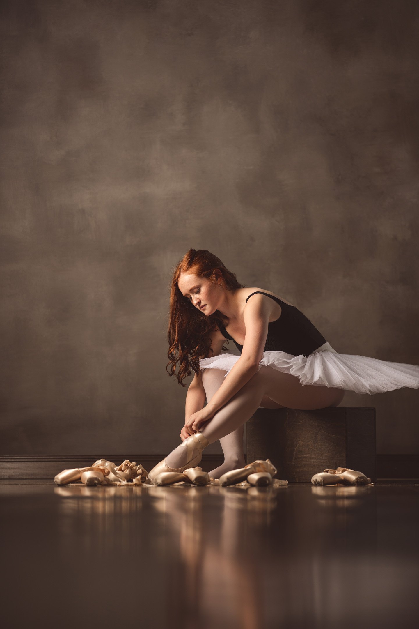 Ballerina lacing up pointe shoe in tutu and black leotard surrounded by ballet shoes