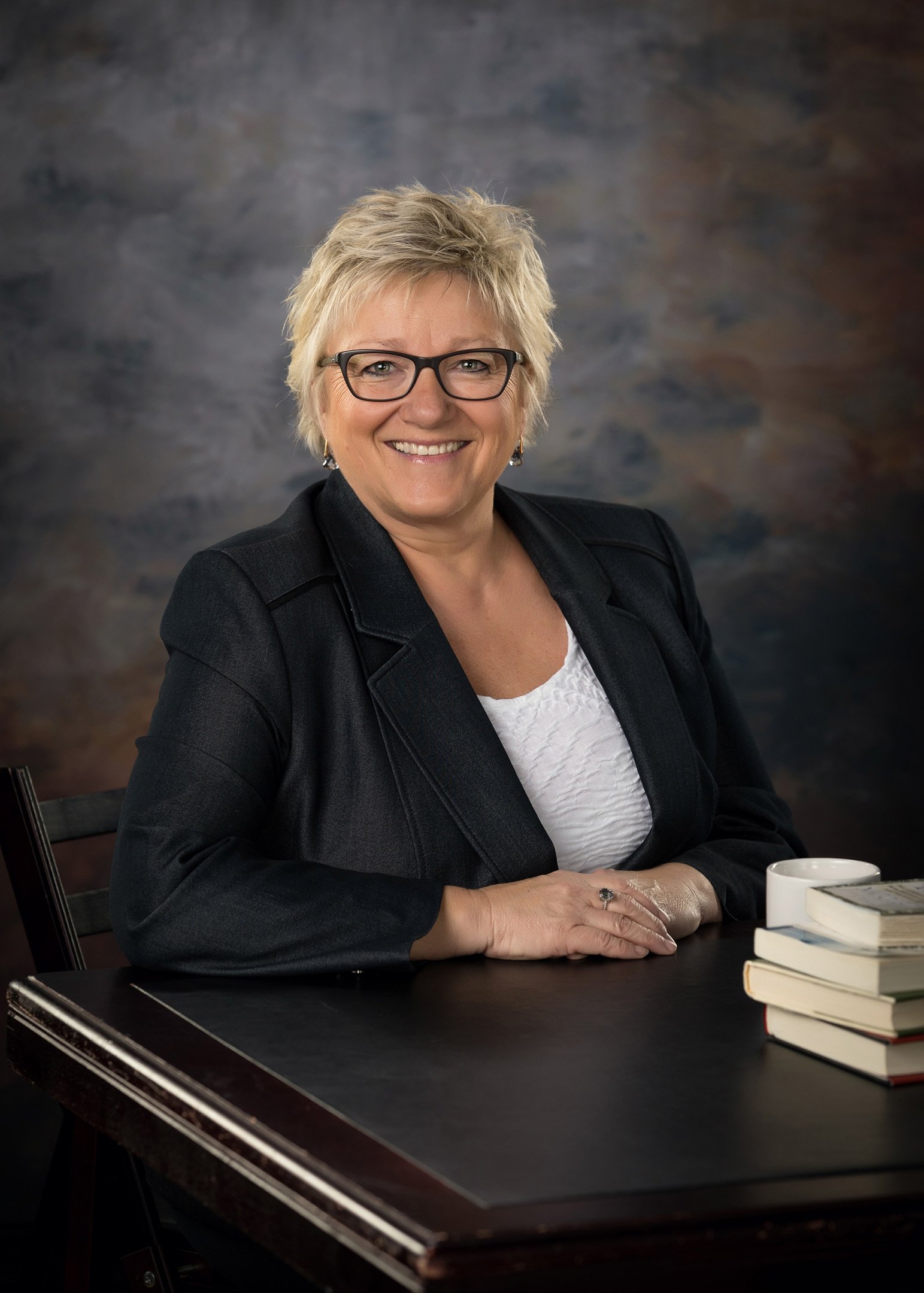 Headshot taken of woman sitting at desk in black suit with books