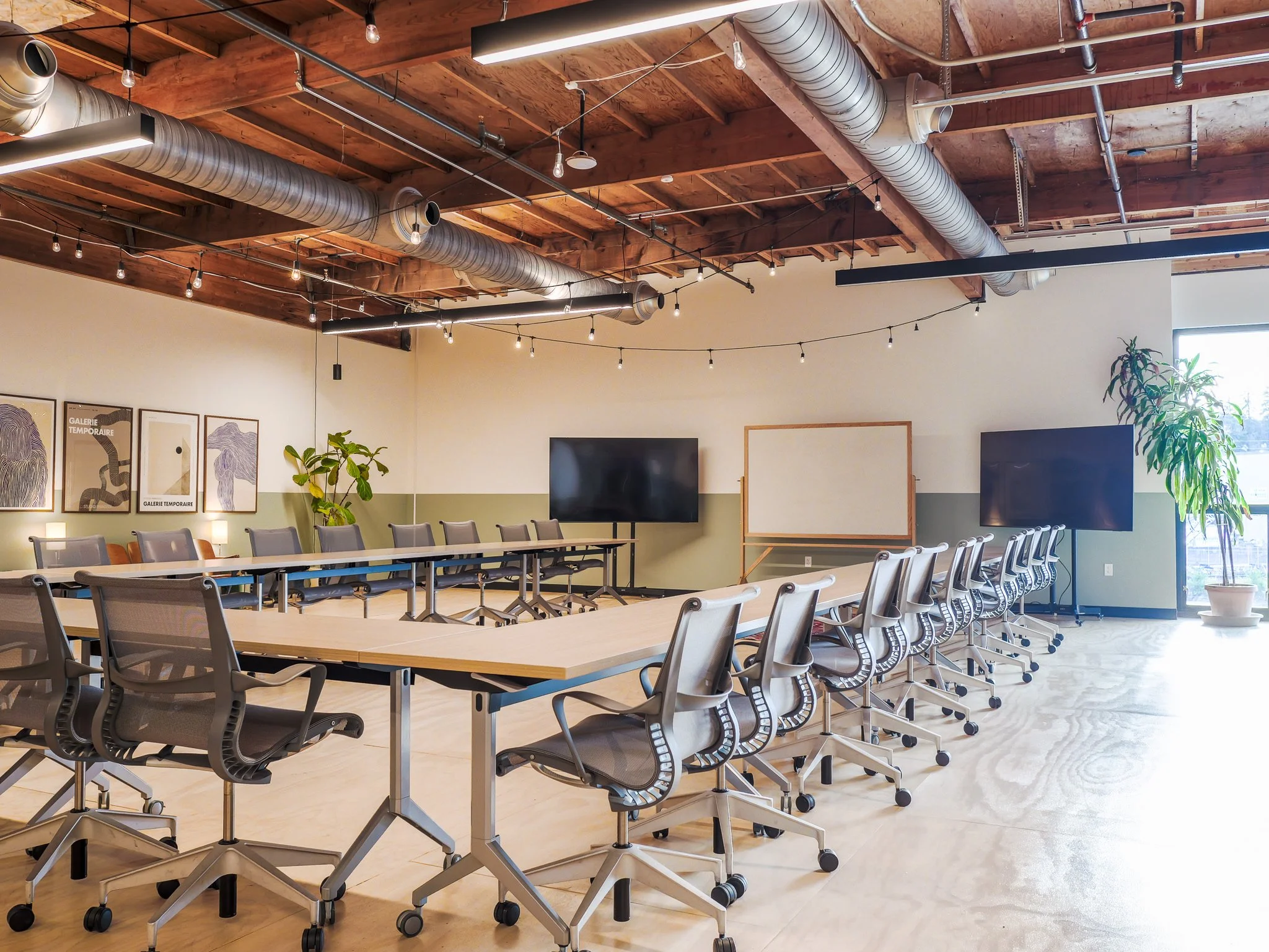 A large U-shaped conference room with a plant and art on the left wall and a whiteboard flanked by two TVs at the front of the room