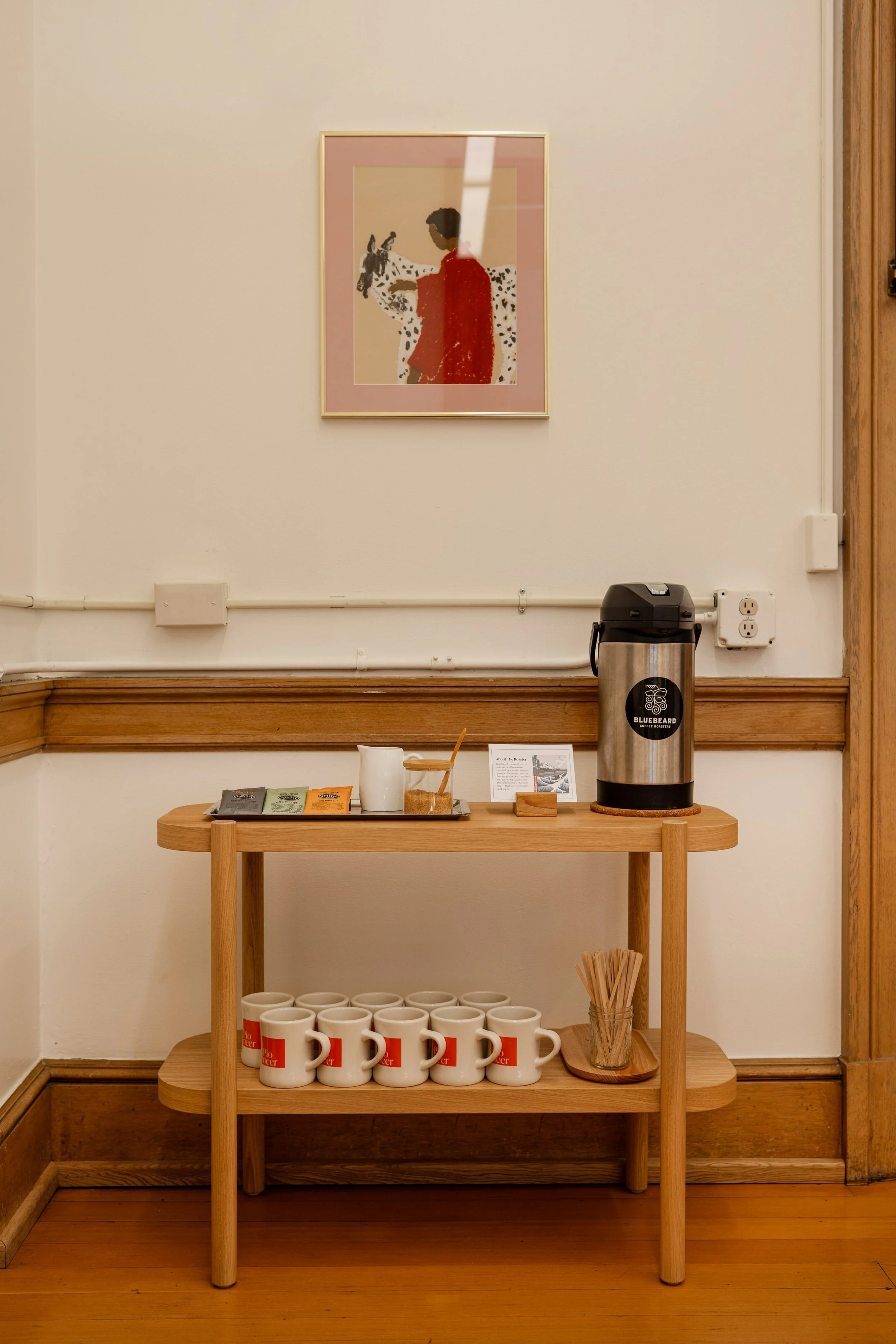 A photo hangs above a small two tier table which holds a coffee carafe, teas, and mugs
