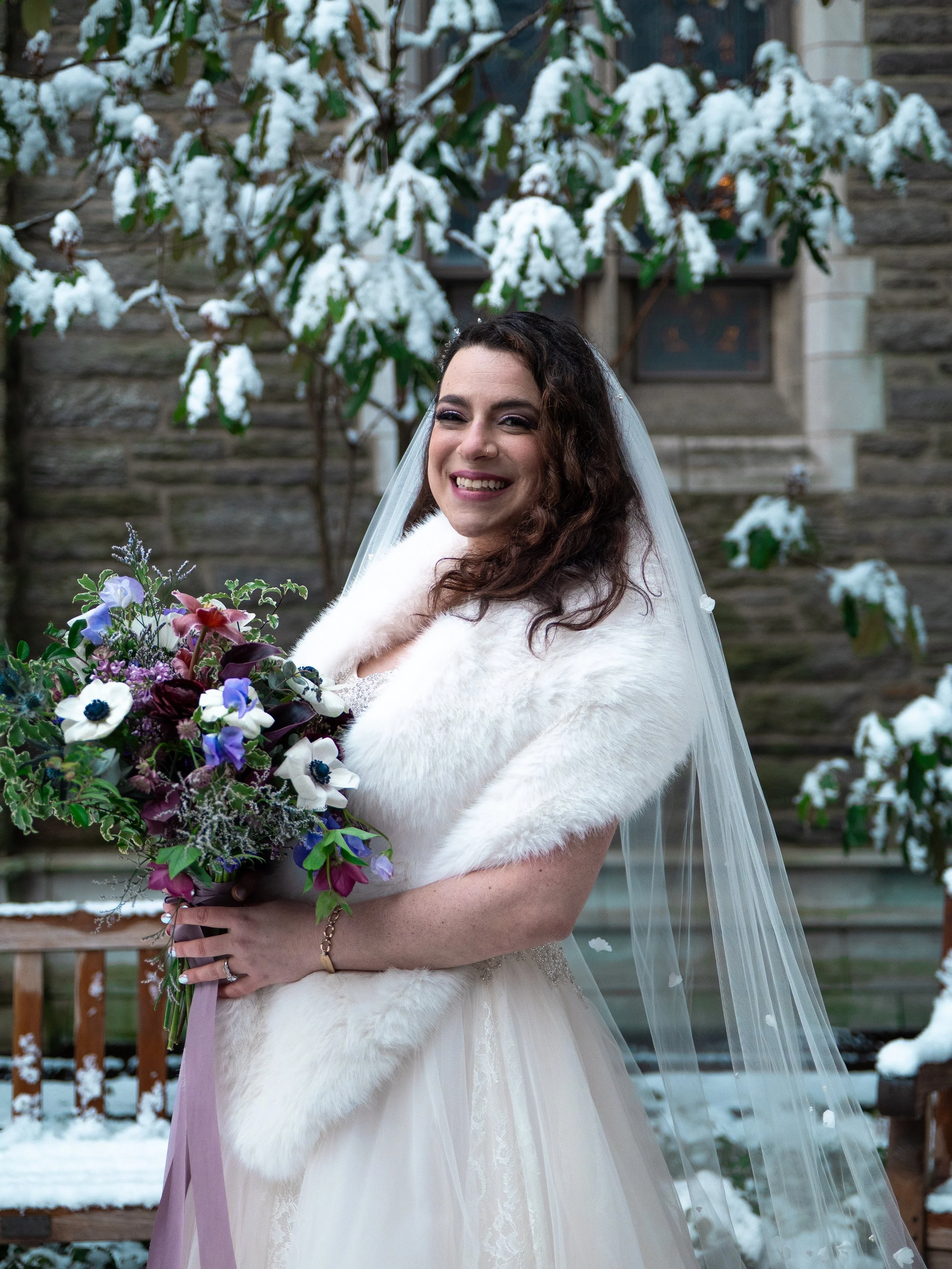 A bride in a white wedding dress with a white fur stole and a long veil holding a bouquet of purple, white, and blue flowers, smiling outdoors in a snowy setting.