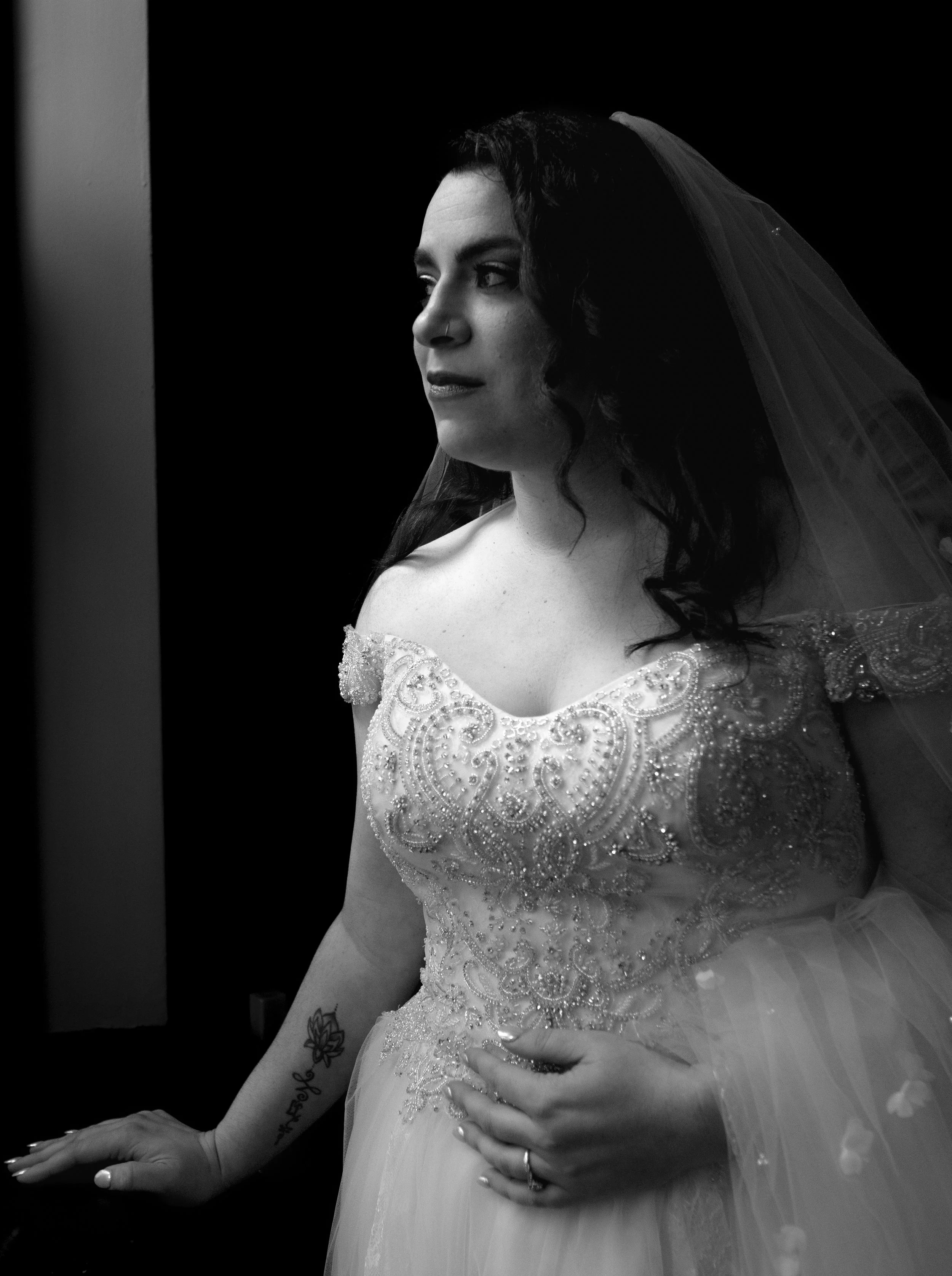 Black and white photo of a woman in a detailed wedding dress, looking slightly to the side, with dark hair, a veil, and a tattoo on her left arm.