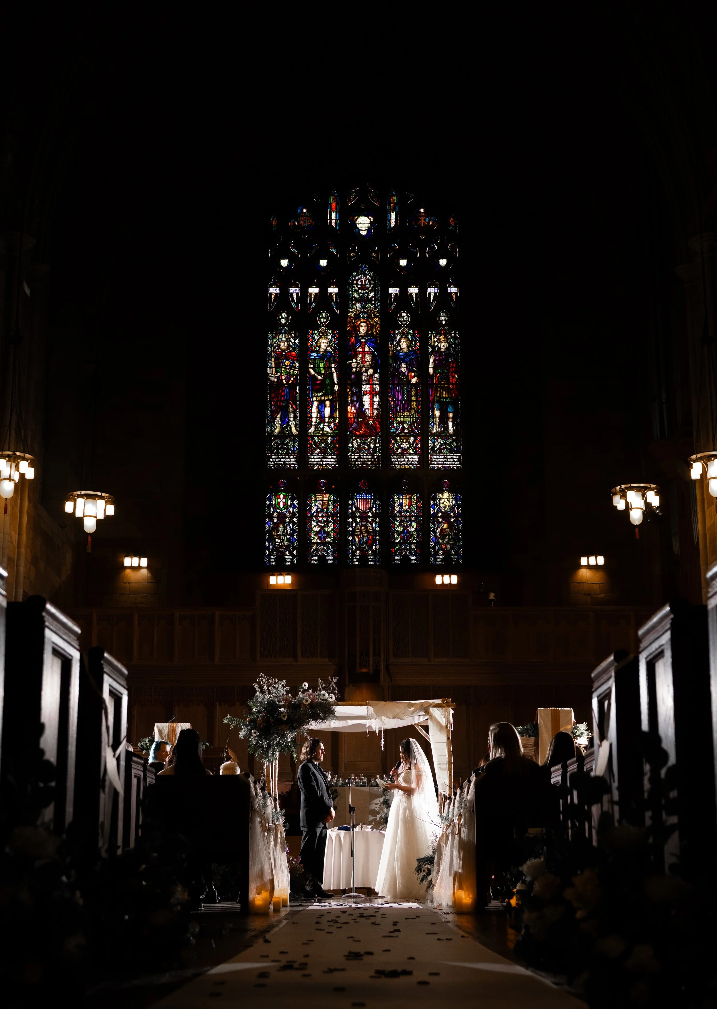 A wedding ceremony taking place inside a church with stained glass windows, with the bride and groom standing before an officiant, surrounded by guests seated in pews.