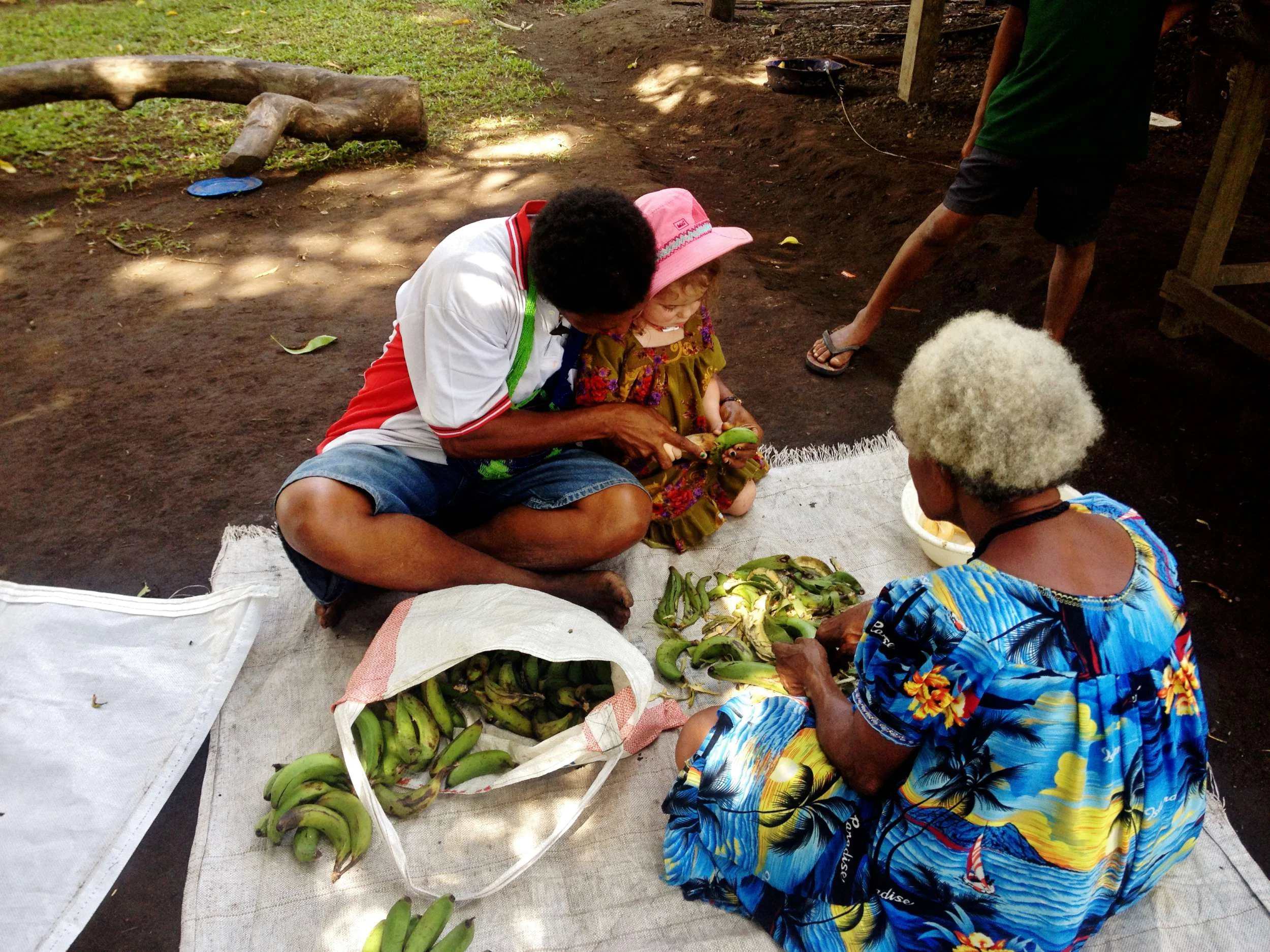 Auntie Suzanne and Auntie Niwe teach Millie how to skin cooking bananas with a pig bone scraper, and cut beans with a super sharp knife, respectively. And Mama stands by to offer encouragement and gumballs at a moments notice.