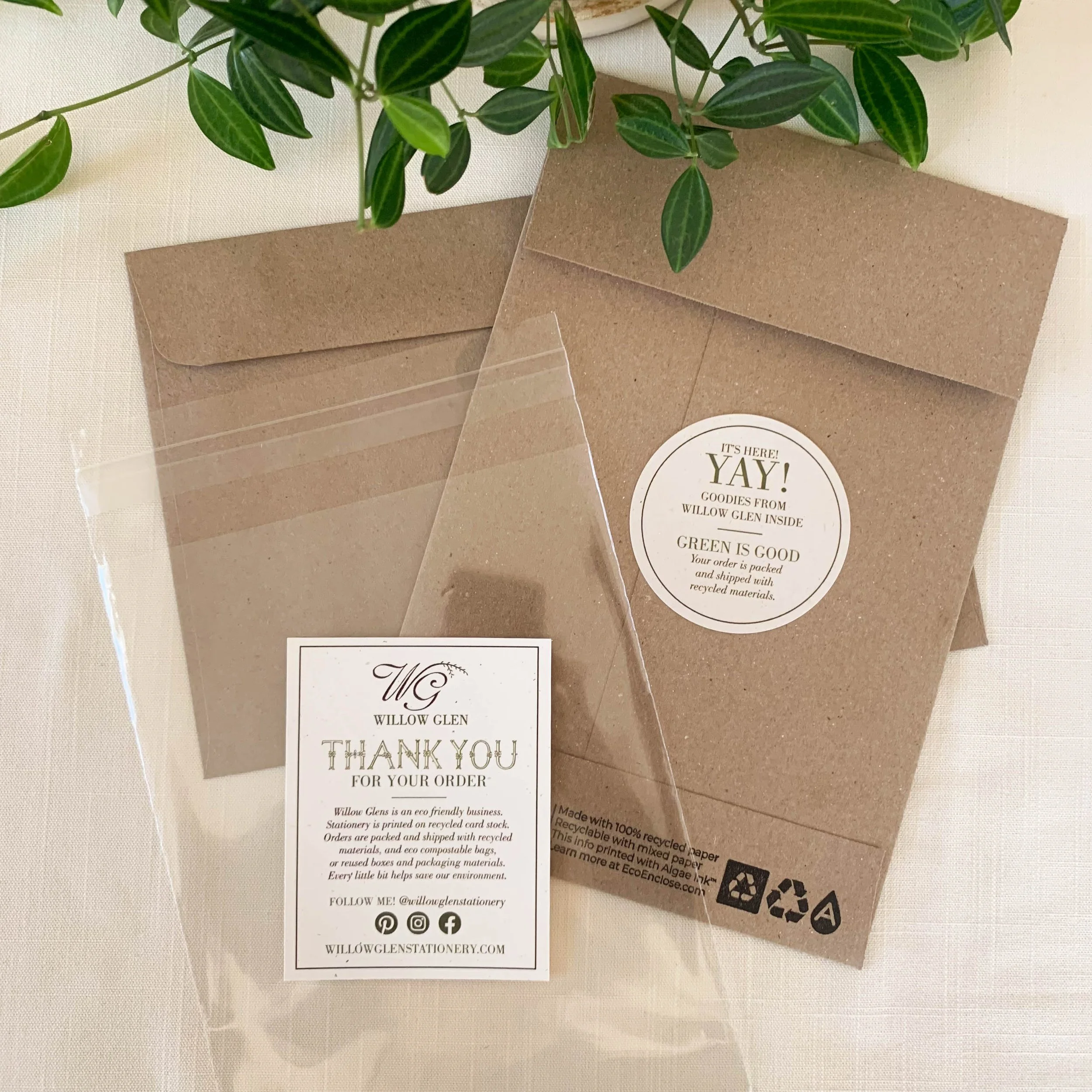 Brown paper envelopes, one with a round white sticker that says 'It's here! Yay! Goodies from Willow Glen inside. Green is good. Your order is packed and shipped with recycled materials.' A separate small, clear plastic bag with a white label that reads 'Willow Glen Thank you for your order. Willow Glen is an eco-friendly business. Stationery printed on recycled paper and shipped with recycled materials. Follow us @willowglentastionery. WillowGlenStationery.com.' There is also a green leafy plant at the top of the image on a light-colored surface.