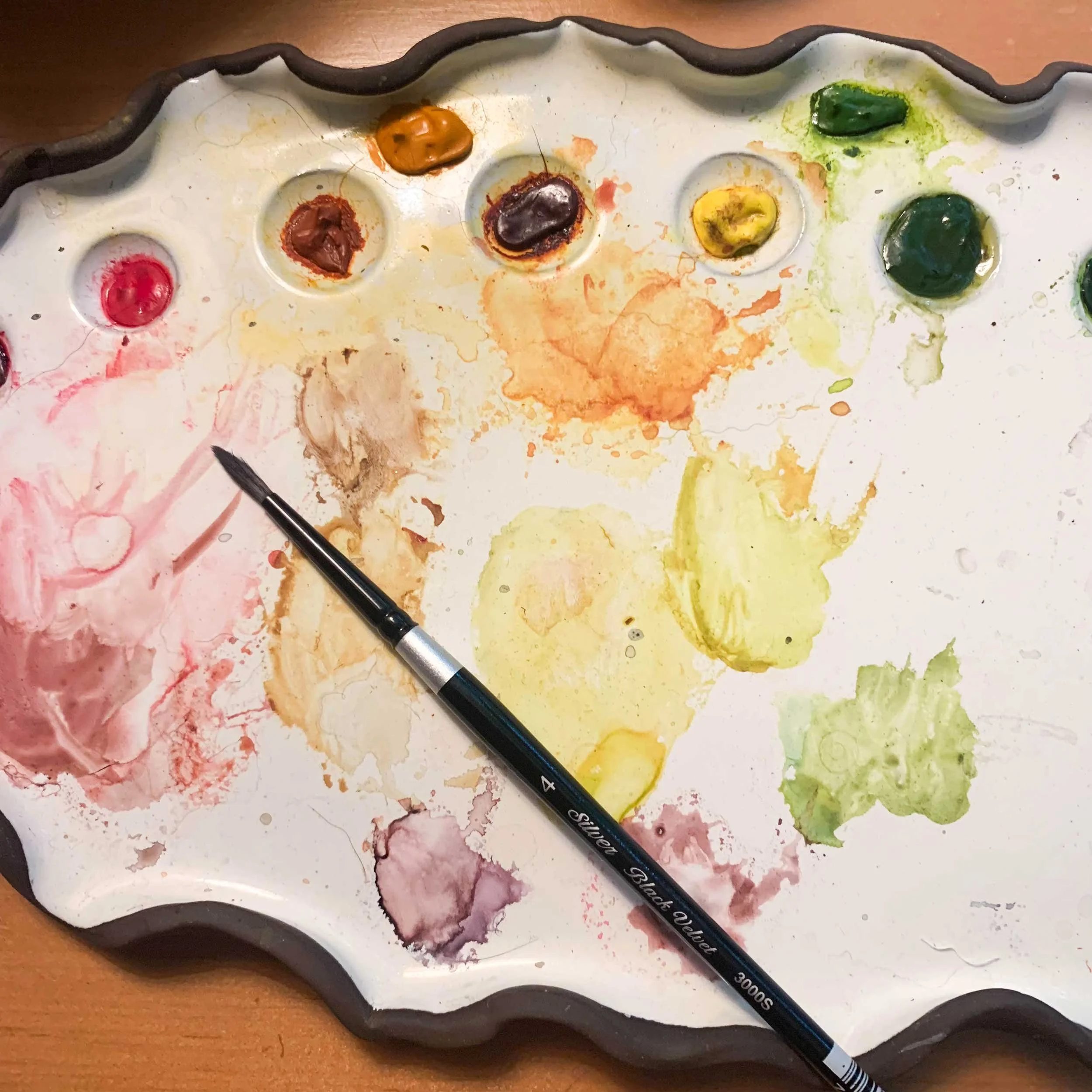 A watercolor palette with various colors, some mixed or spilled, and a paintbrush resting on it.