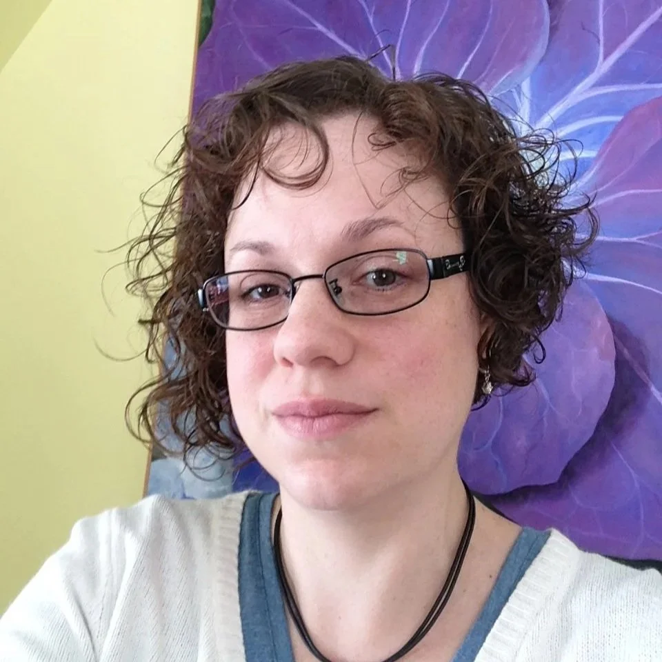 A woman with curly brown hair and glasses taking a selfie in front of a purple flower painting.