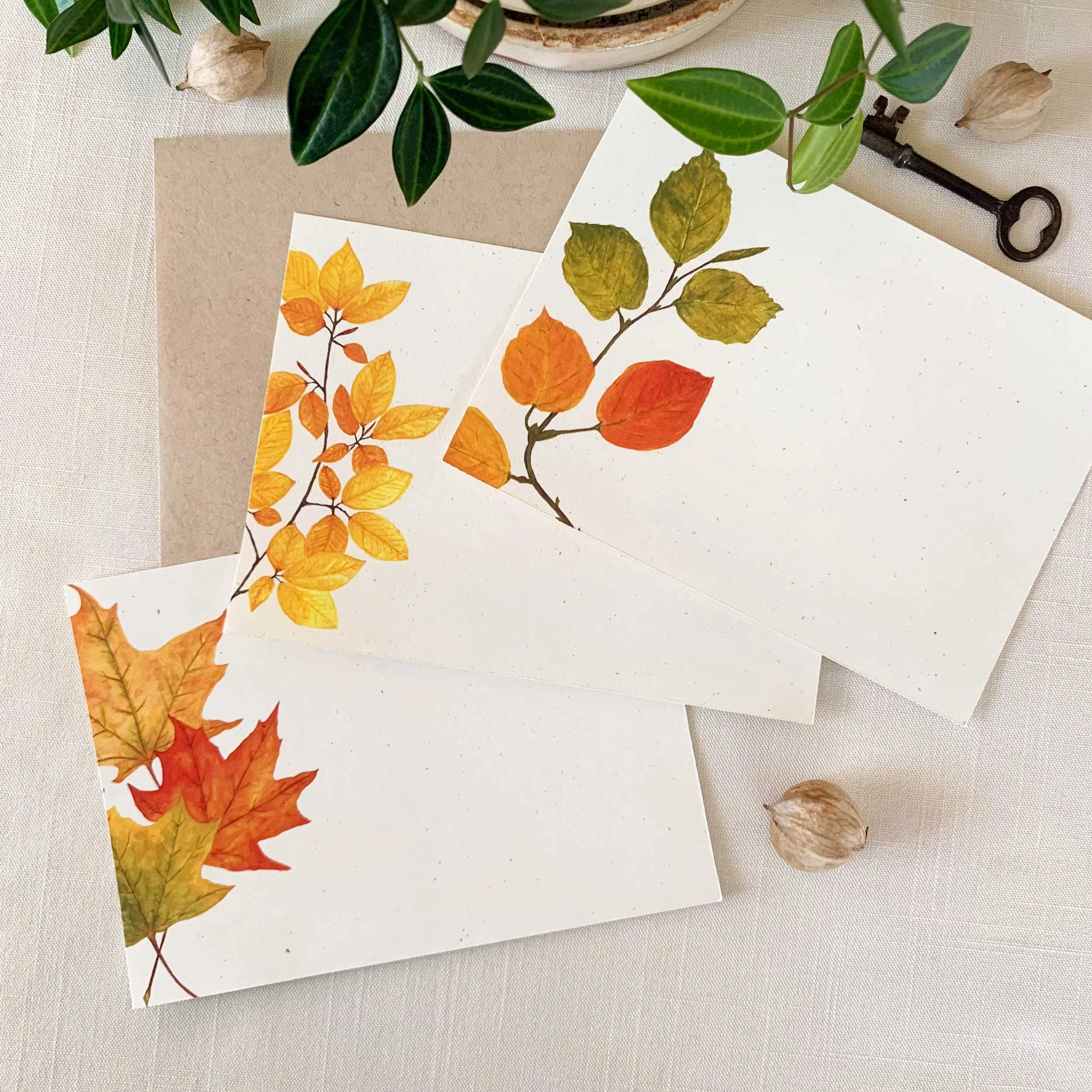 Three pieces of paper with fall leaves illustrations in orange, yellow, and red hues. Decorations include a potted plant, an old-fashioned key, and dried seed pods on a white surface.