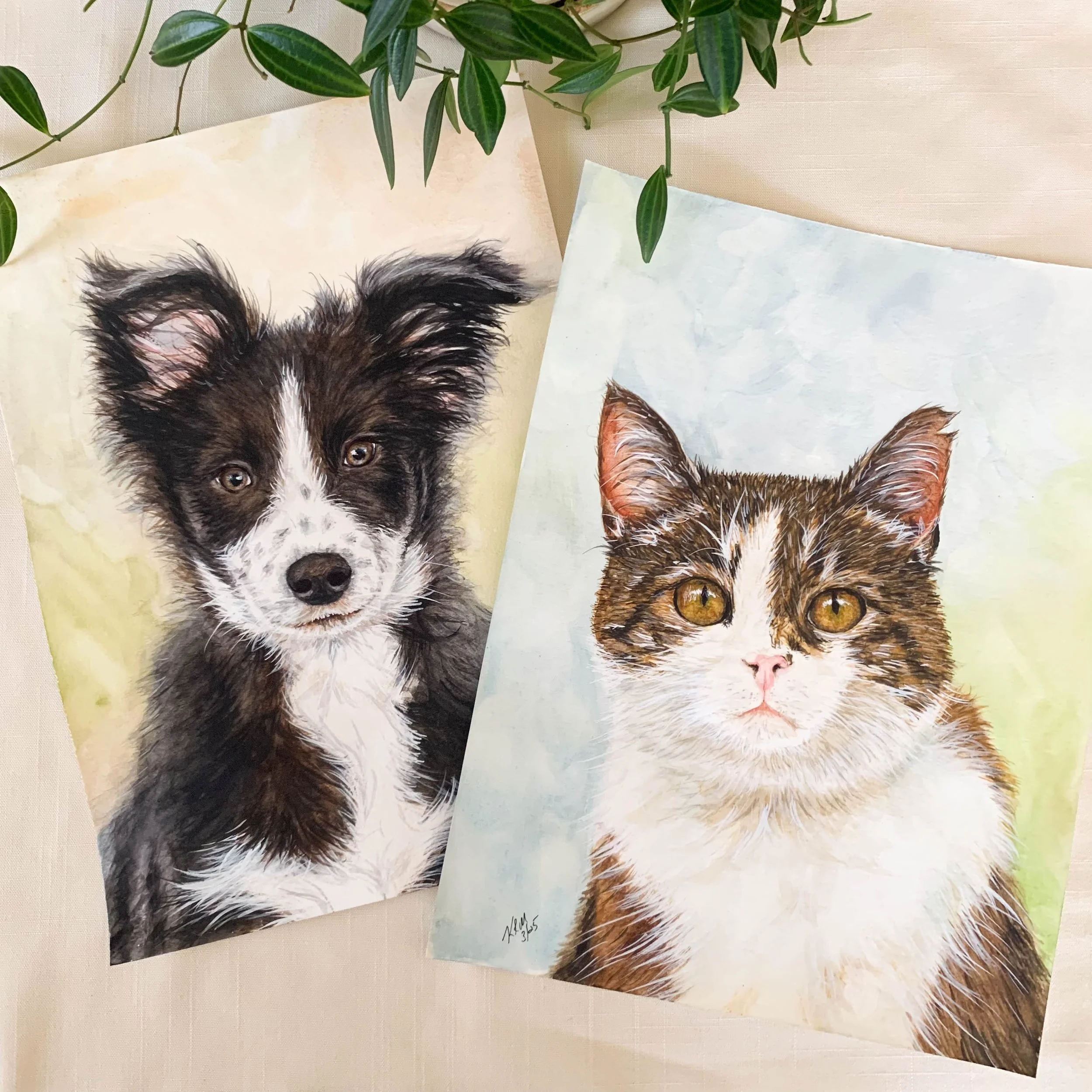 Two watercolor paintings of animals laid on a beige surface with green leaves in the background. The left painting depicts a black and white dog with large upright ears and expressive eyes. The right painting shows a brown and white tabby cat with yellow eyes and pointed ears.