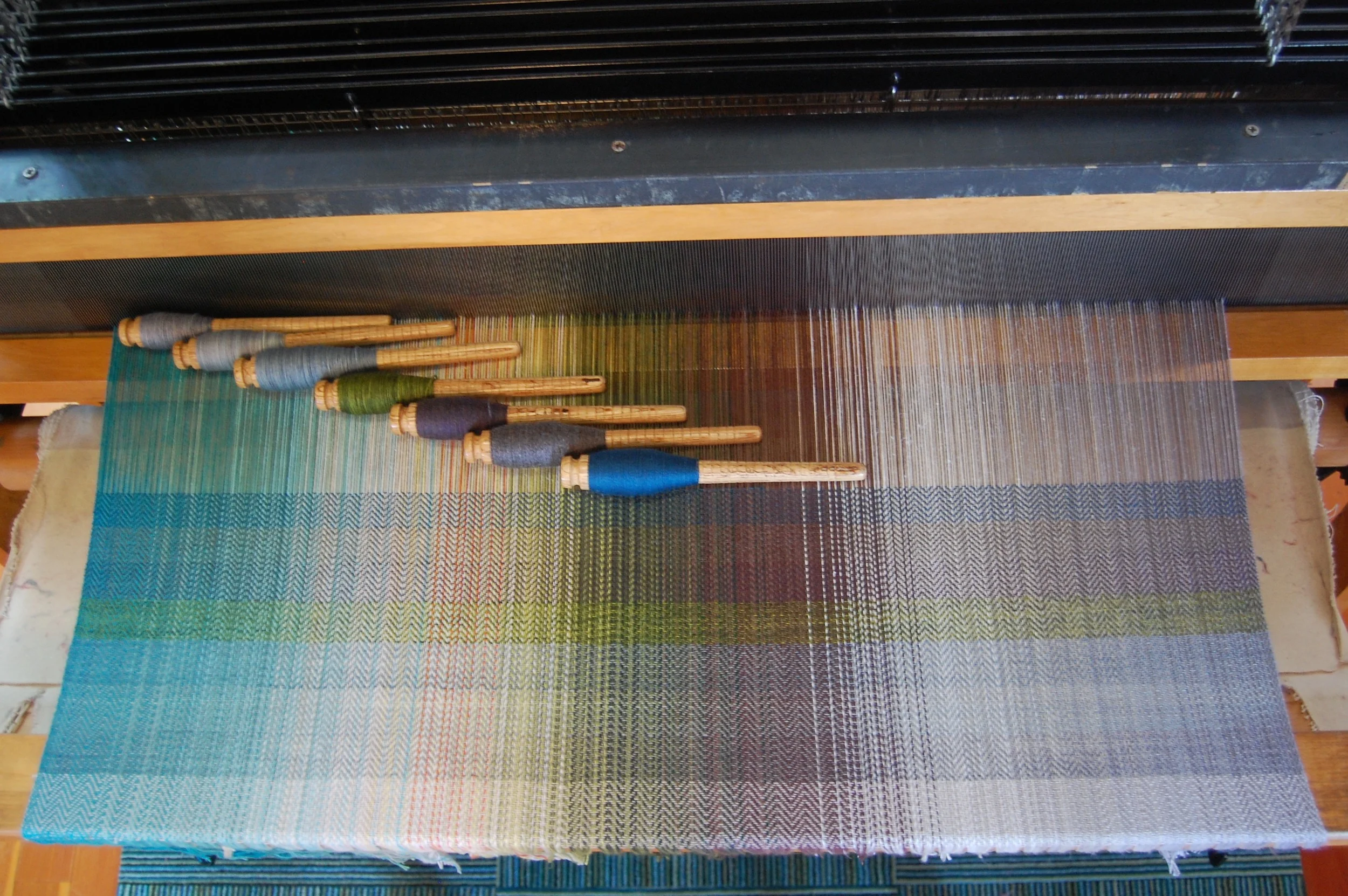 Superwash wool wefts - from bottom of woven sample and left of pirns (lowest color is the silver cotton): MadTosh Superwash merino lace in Aura, Mica, Well Water and Jade; Superwash merino lambswool in Midnight, Steel and Lapis.