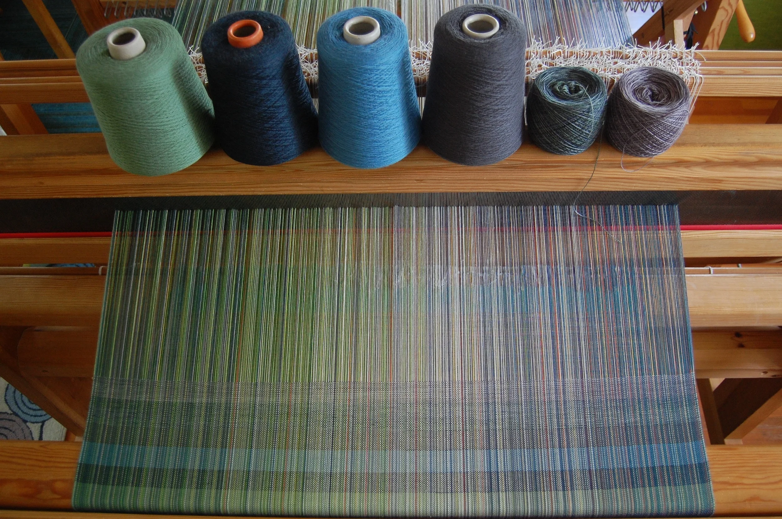 The wools, from the bottom of the woven sample (and from the left of the cones): tourmaline superwash merino lambswool, mallard superwash merino lambswool, mood blue organic merino wool, charcoal organic merino wool, MadTosh Superwash Lace in Fir Wr…