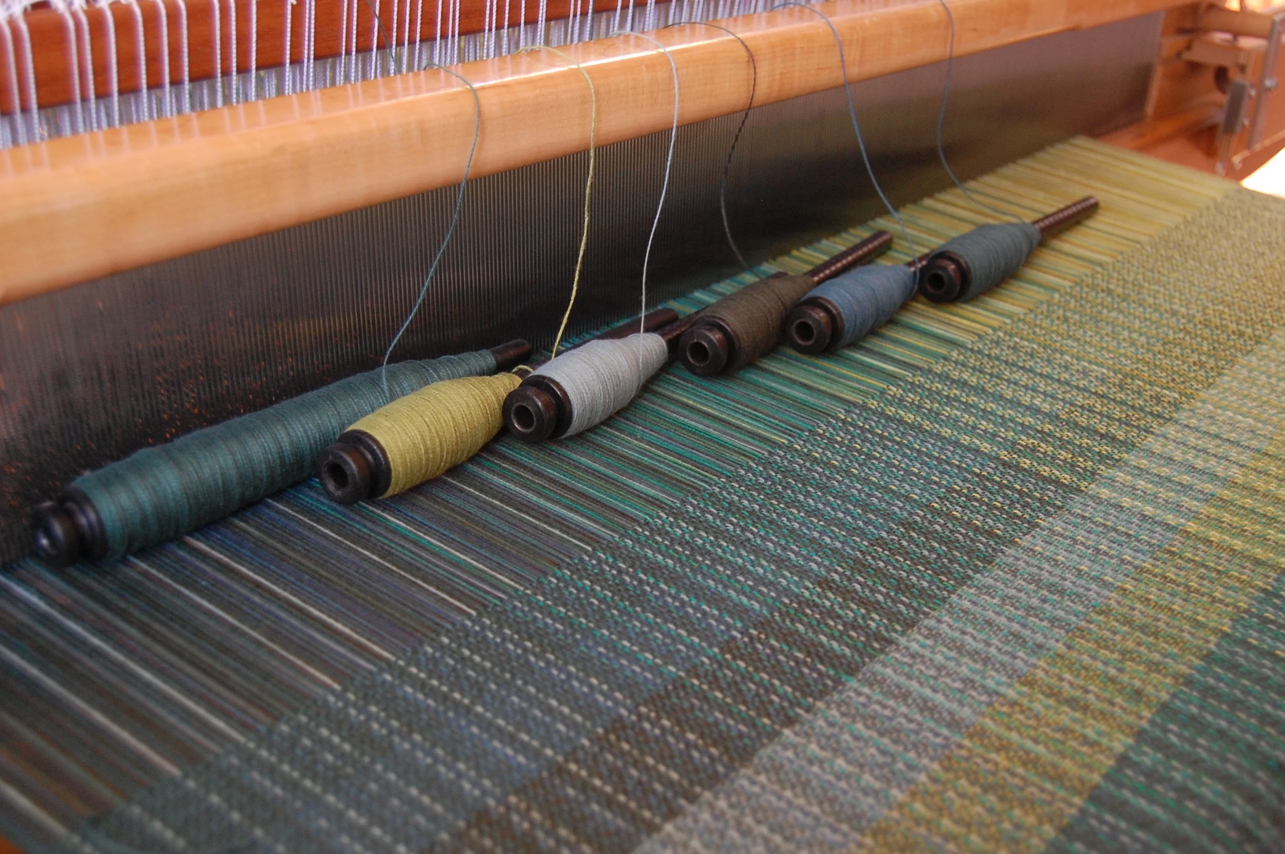 weft testing with cotton. from the bottom of the woven sample and the left of the pirn: greenish-teal EC, fern green organic cotton, light teal cotton, forest green, blue-ish teal EC, peacock organic cotton.