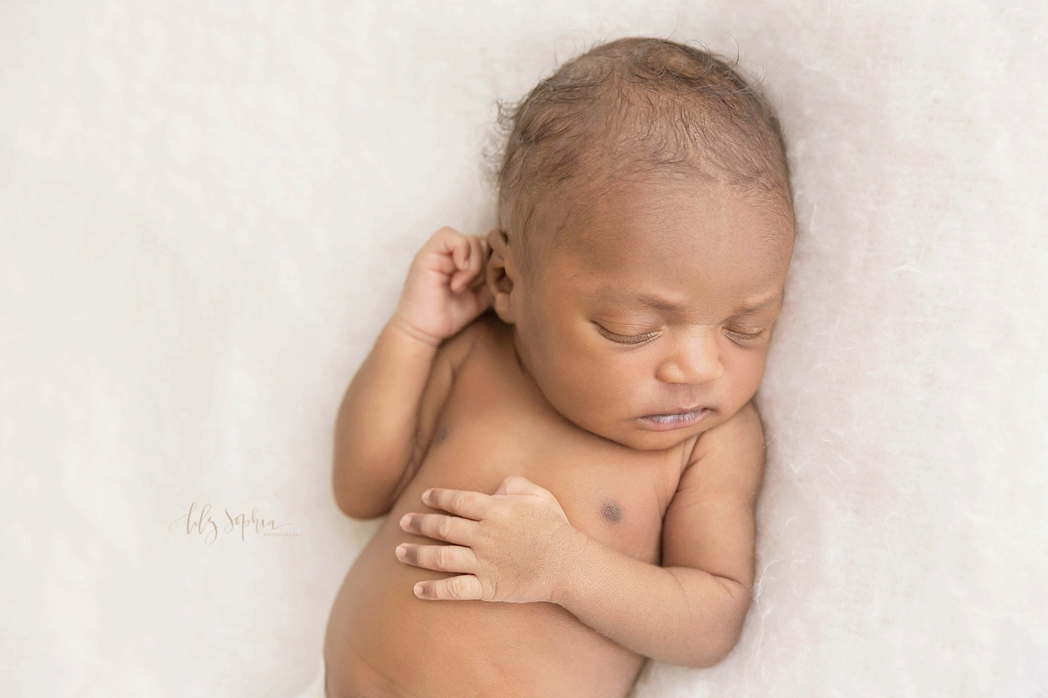  Image of a sleeping African American newborn on a white blanket, with one hand on her belly and the other hand by her ear.&nbsp; 