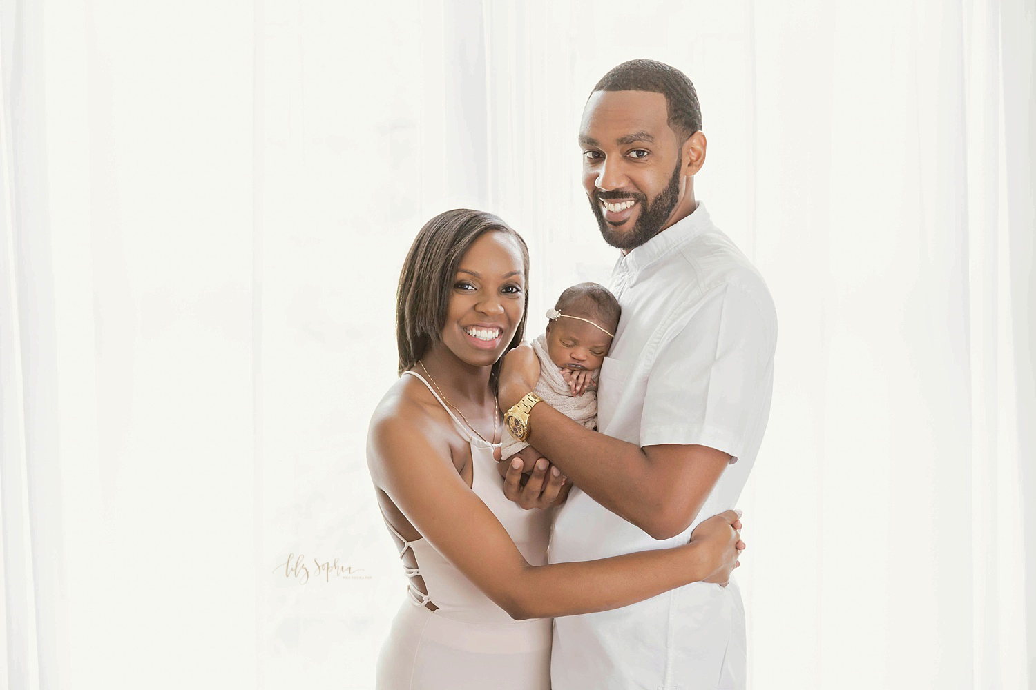  Image of a African American family of three. The father is holding his sleeping, newborn daughter and smiling at the camera while his wife has her arms around his waist while also smiling at the camera.&nbsp; 