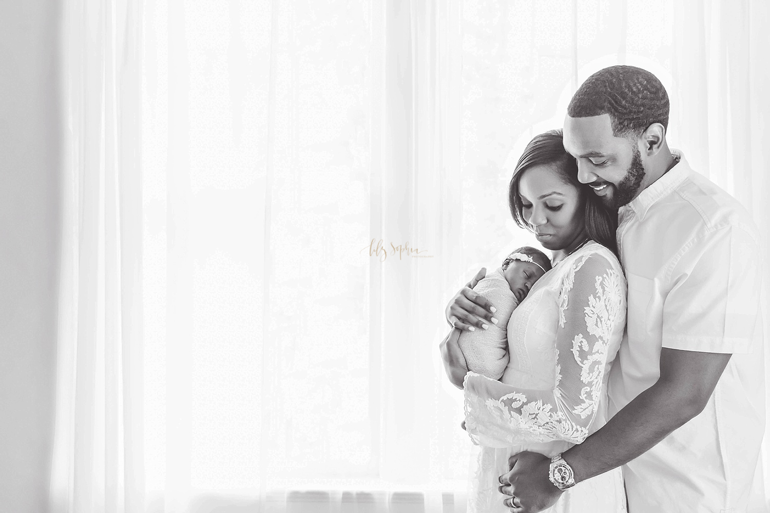  Black and white image of an African American family. The mother is holding her sleeping newborn daughter, while the father stands behind her, smiling down at them both, with his arms around his wife.&nbsp; 